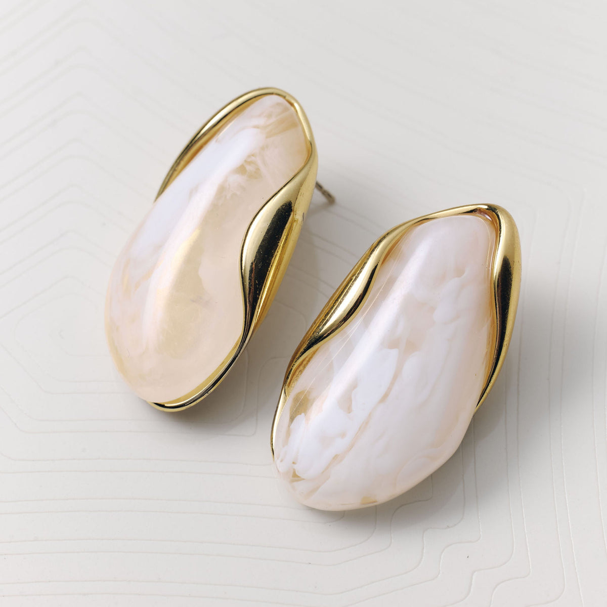 A pair of gold-tone stud earrings with organic, wavy teardrop-shaped cream and white marbled stone inlays, displayed on a light beige textured surface.