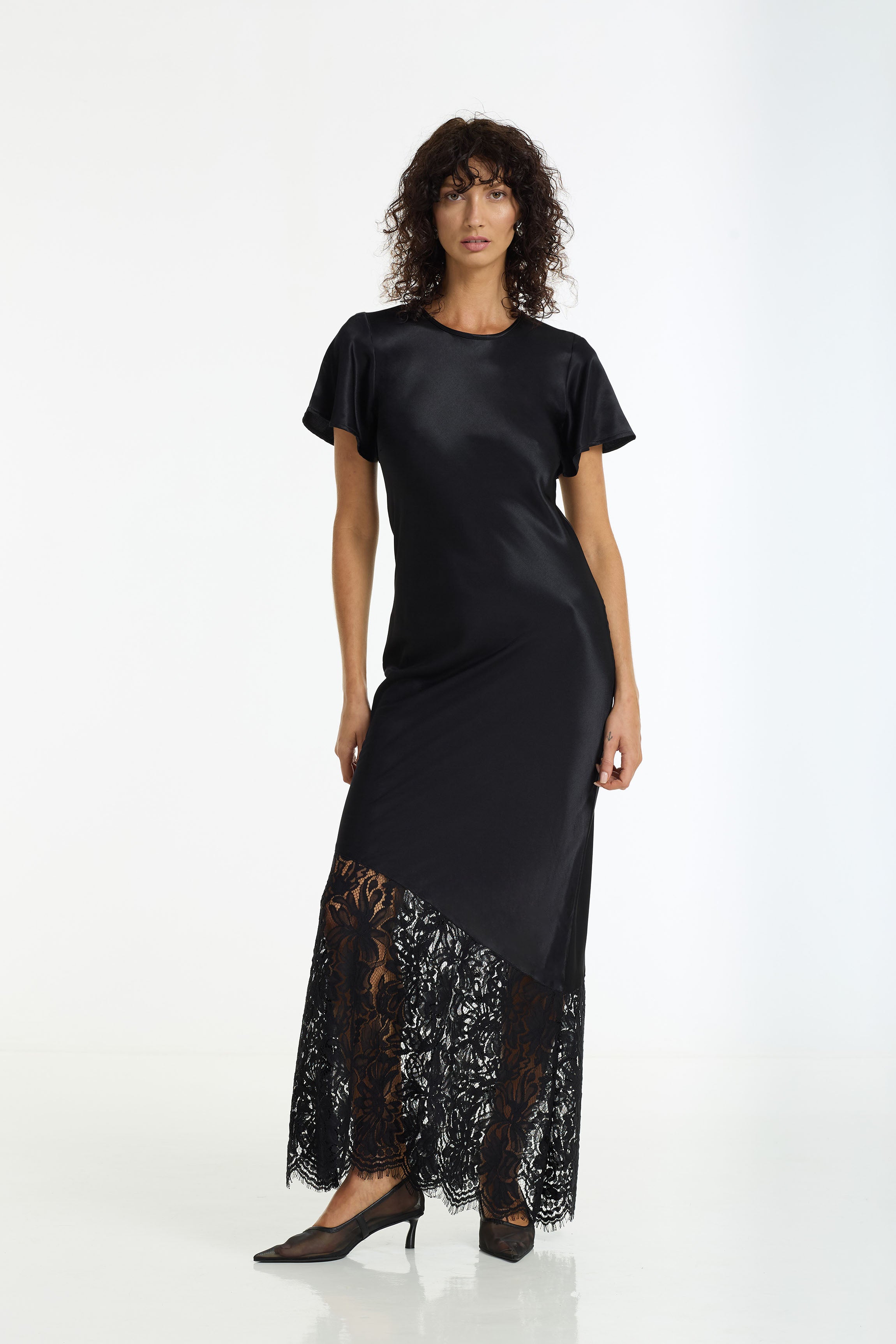 A model wearing a After Dark Satin Lace Maxi Dress in black by BLAK, paired with black pointed-toe heels, against a plain white studio background.