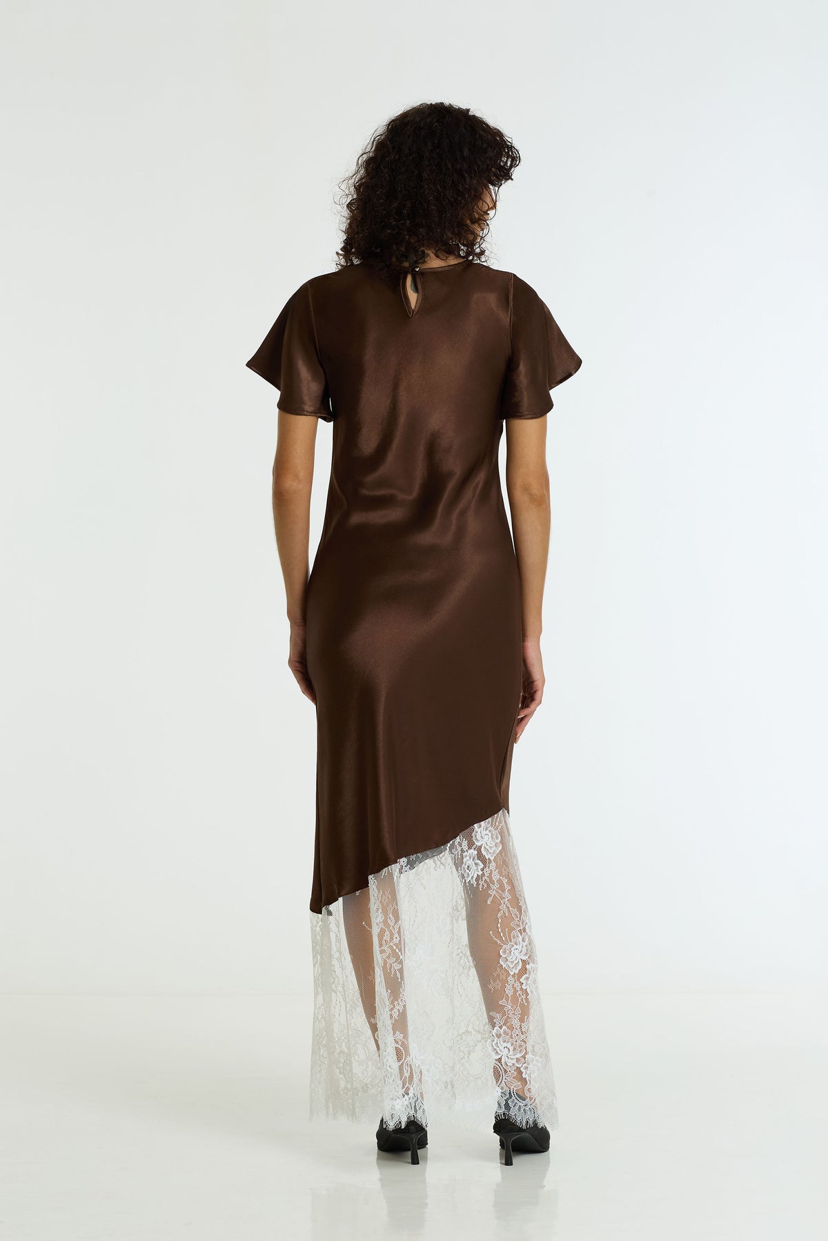 A woman viewed from behind wearing a brown satin dress with flutter sleeves and a sheer white lace overlay at the hem, photographed against a plain white studio background.