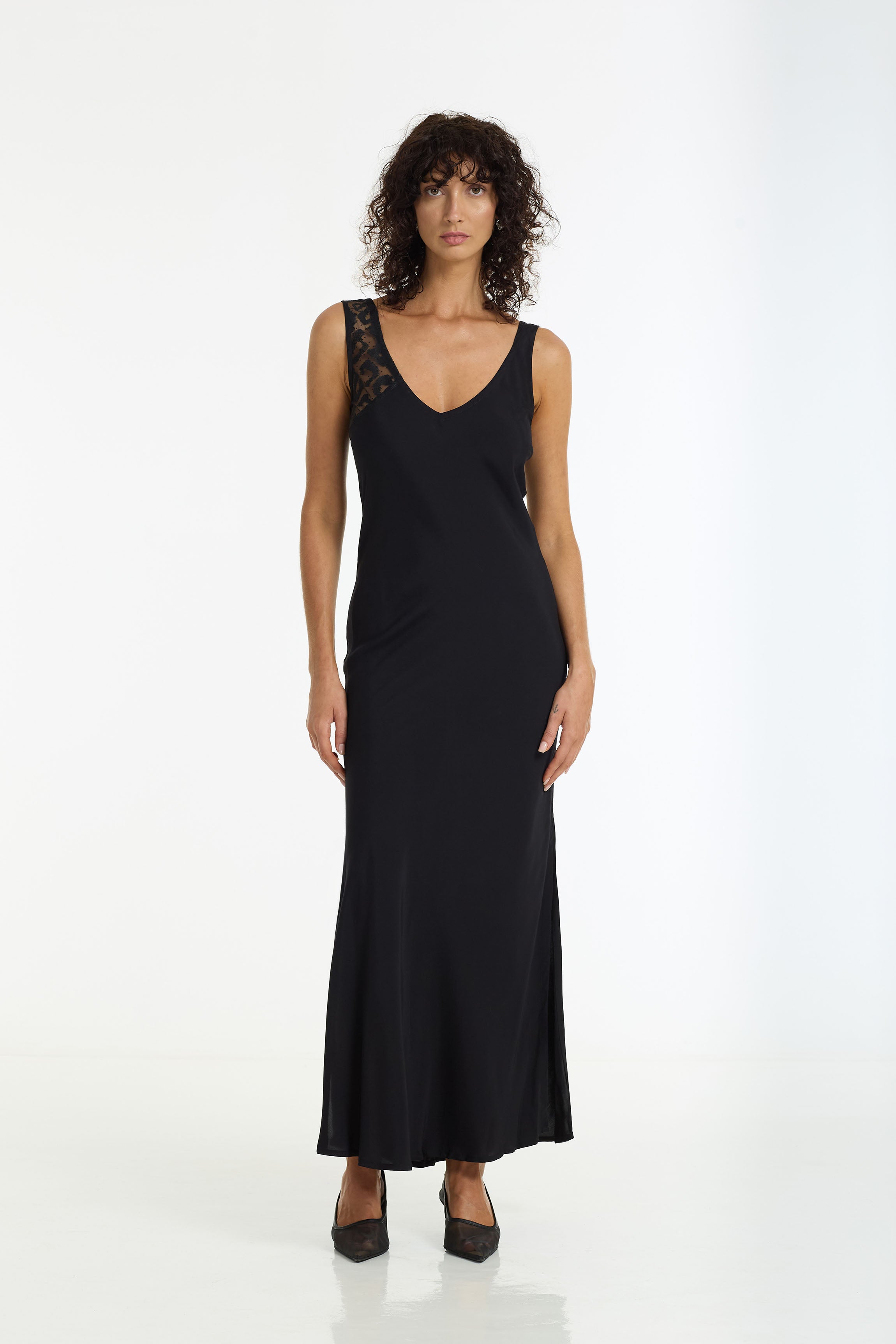 A woman modeling a Alex Bias Cut Maxi Dress in black by BLAK with lace detailing on the upper left shoulder, standing against a plain white studio background.