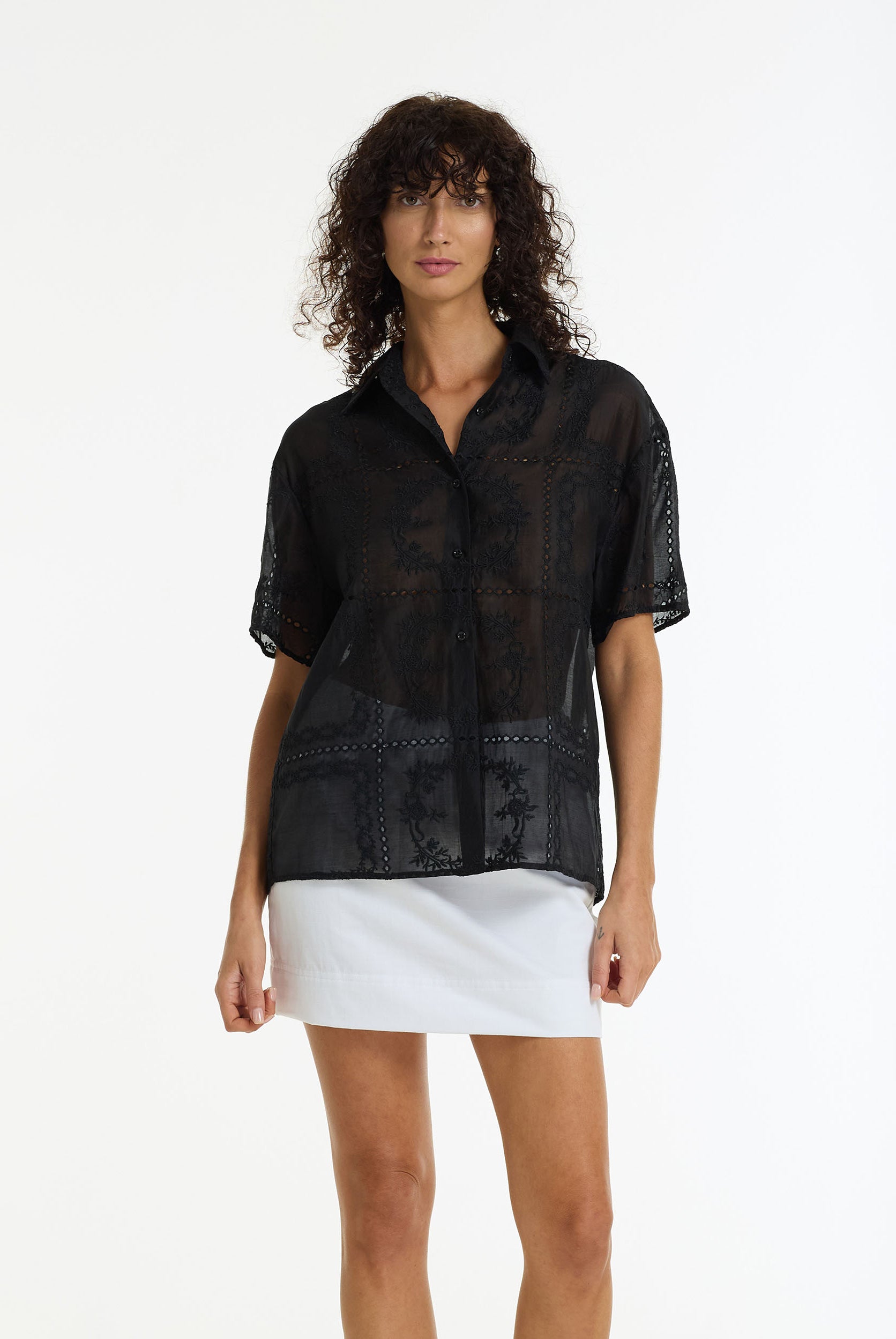 A model wearing the Align Short Sleeve Anglaise Shirt black by BLAK over a white mini skirt, shot against a plain white studio background.