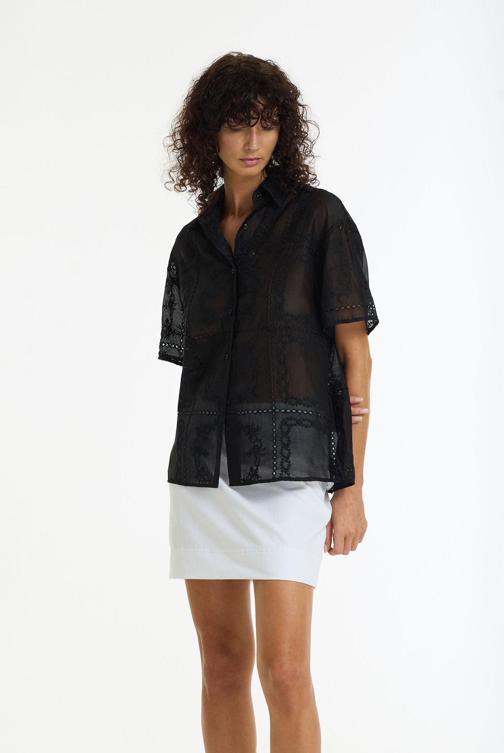 A model wearing the Align Short Sleeve Anglaise Shirt black by BLAK, paired with a white mini skirt, against a plain white studio background.