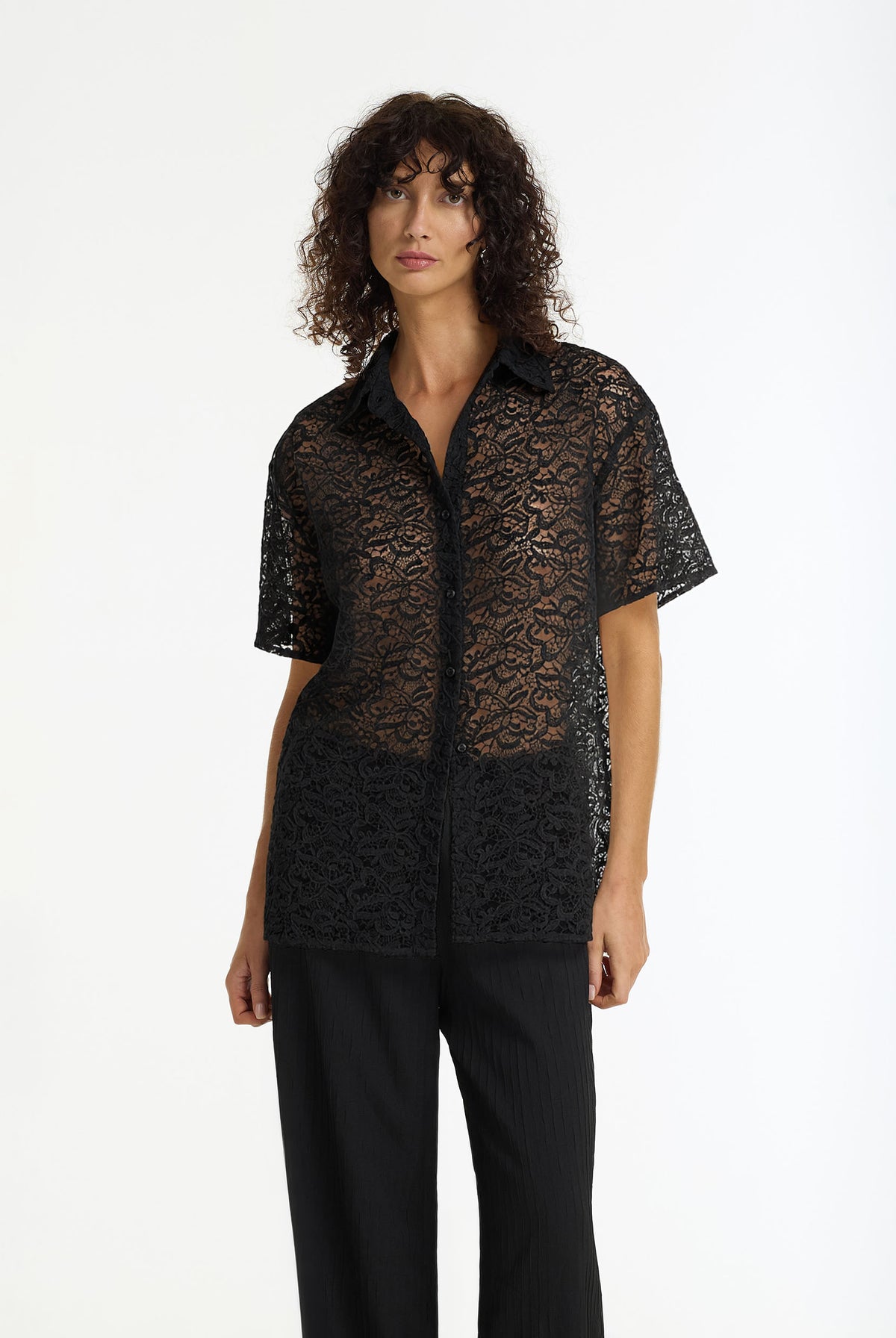 A model with curly dark hair wearing the Align Short Sleeve Lace Shirt black by BLAK and black trousers, photographed against a plain white studio background.