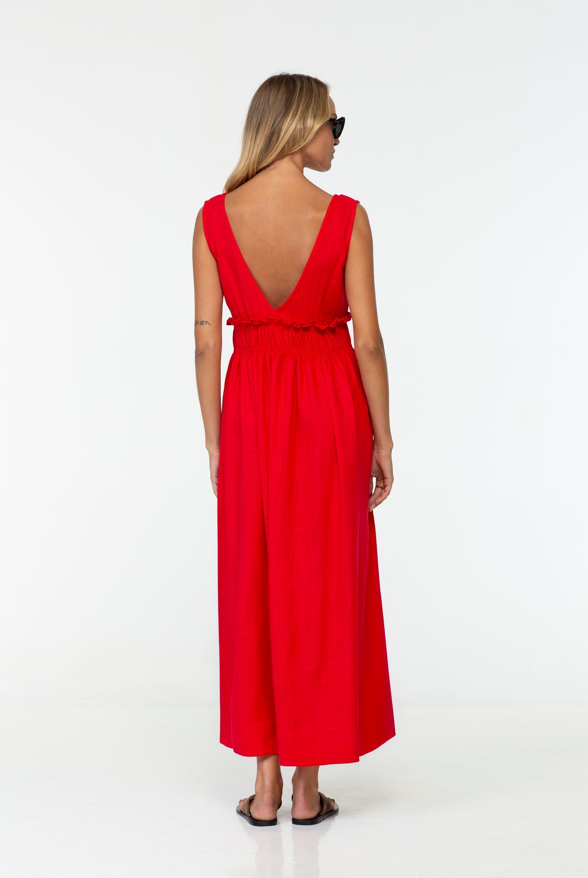 A woman wearing a Anya Linen Maxi Dress in Strawberry Red | BLAK.  It's a bright red sleeveless maxi dress with a deep V-back and smocked waist, shown from the back against a plain white studio background.