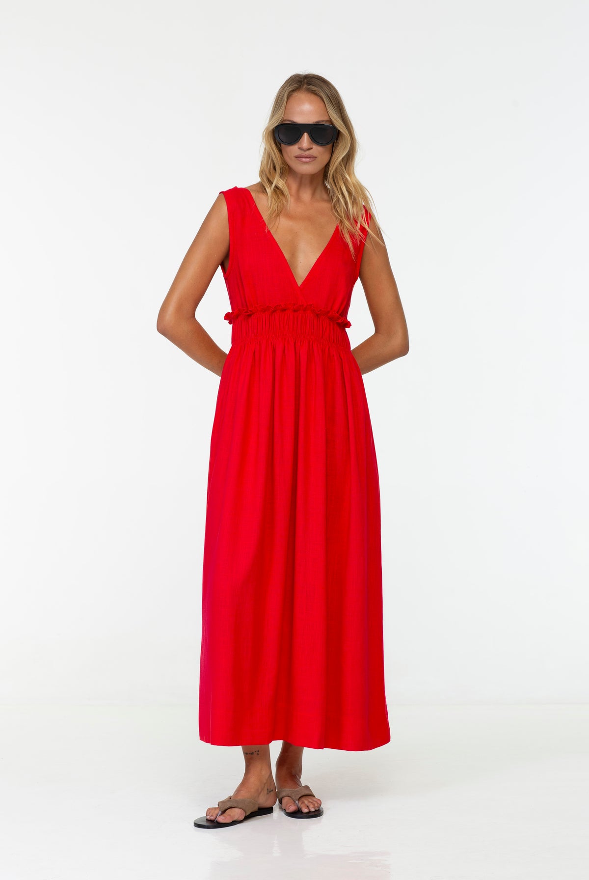 A woman modeling the Anya Linen Maxi Dress Strawberry Red by BLAK. It's a bright red sleeveless maxi dress with a deep V-neckline and a smocked waist, styled with black sunglasses and flat sandals against a plain white studio background.
