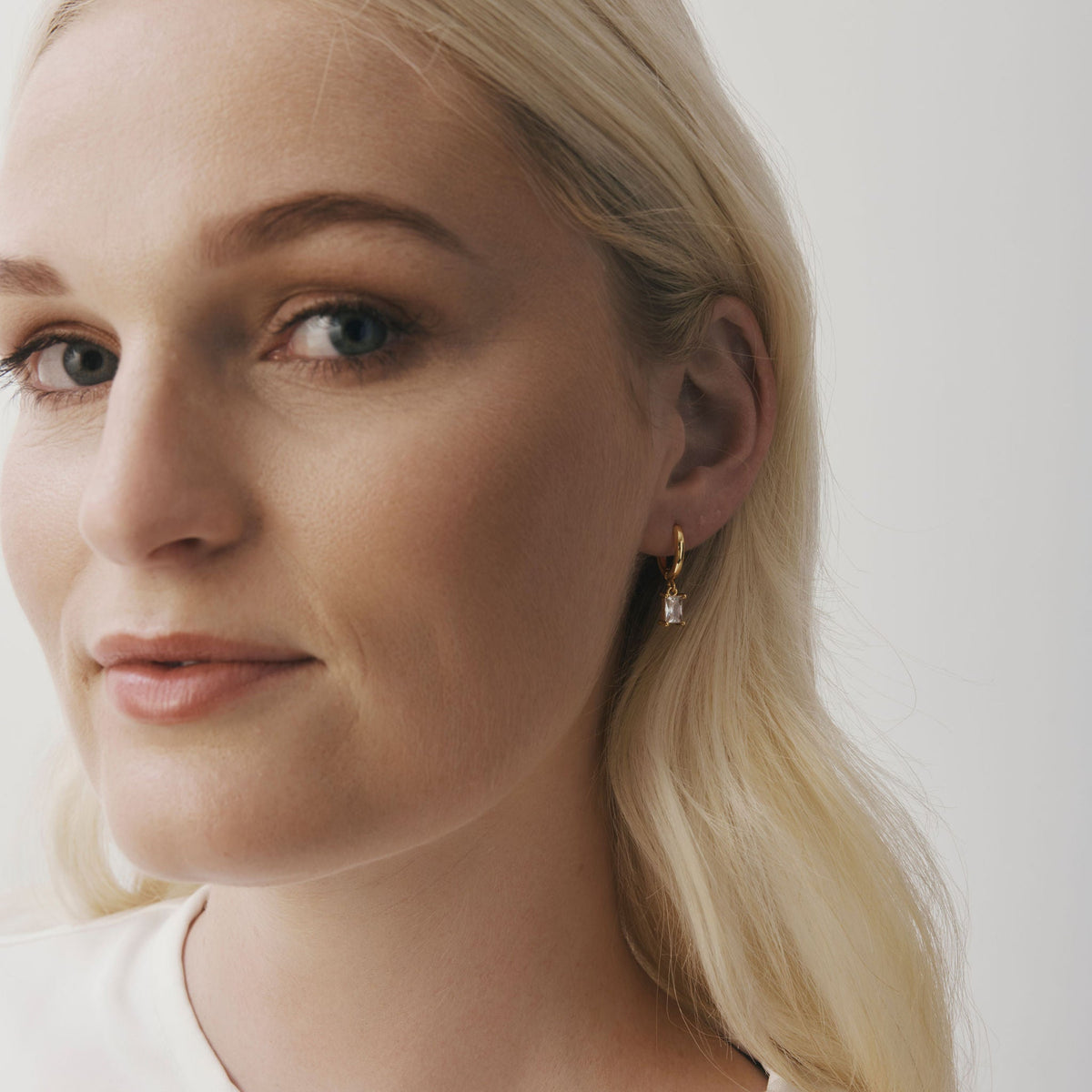 A close-up portrait of a blonde woman wearing a delicate gold hoop earring with a small clear stone, against a plain light background.