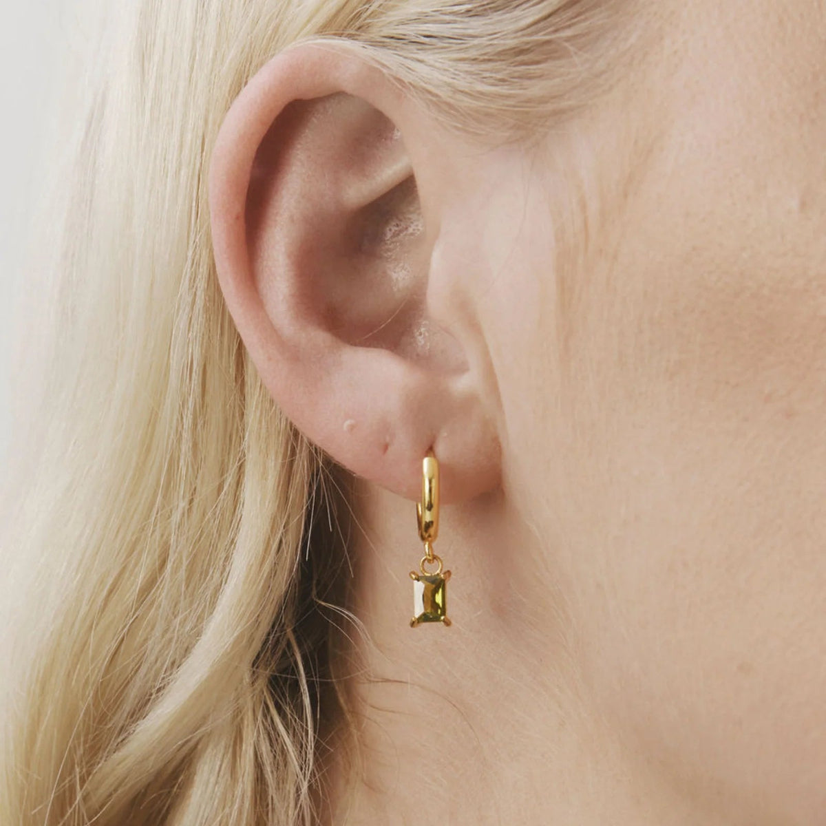 A close-up of a model's ear wearing a delicate gold huggie hoop earring with a small gold hourglass charm.