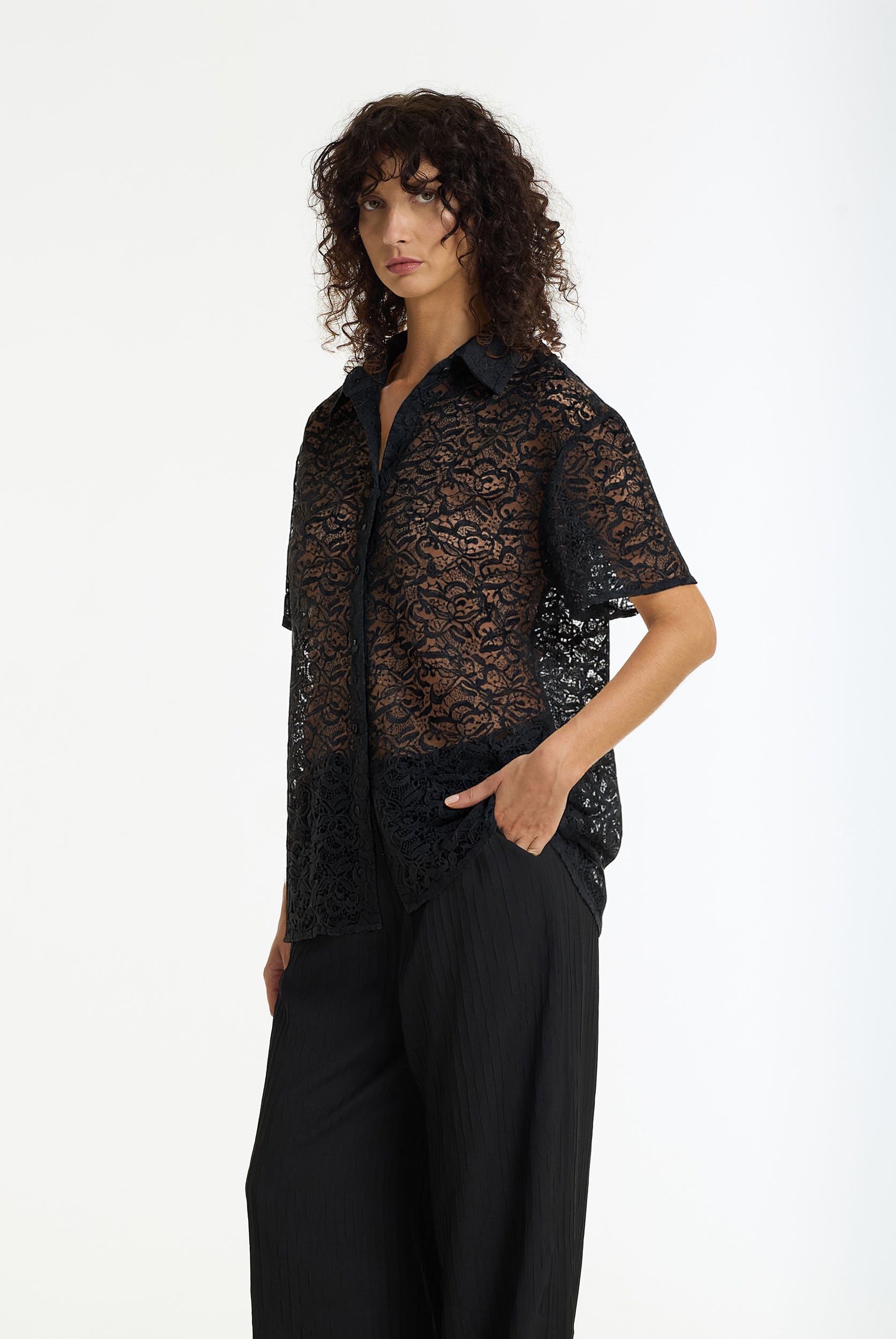 A side view of model with curly dark hair wearing the Align Short Sleeve Lace Shirt black by BLAK and black trousers, photographed against a plain white studio background.