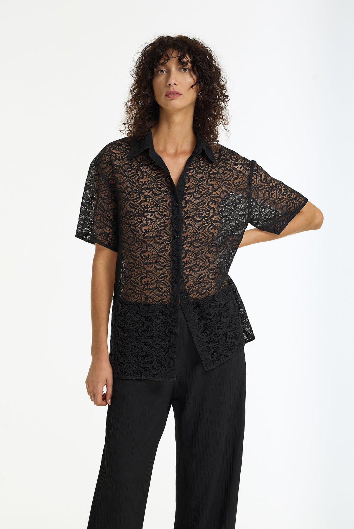 A model wearing a sheer black lace short-sleeve button-up shirt and black wide-leg trousers, photographed against a clean white studio background.