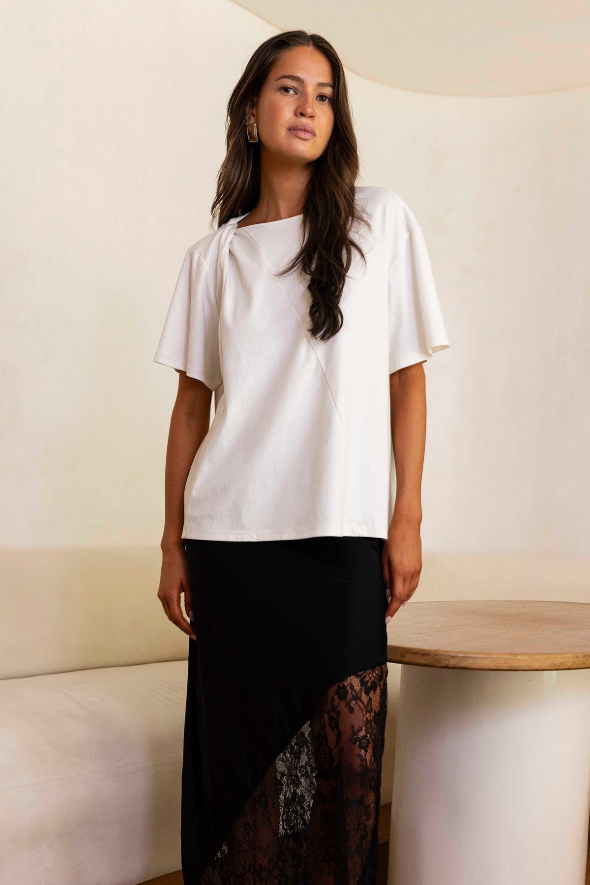 A woman stands in a minimalist room wearing a loose white short-sleeve top and a long black skirt with a sheer lace overlay panel, paired with dark flat sandals.