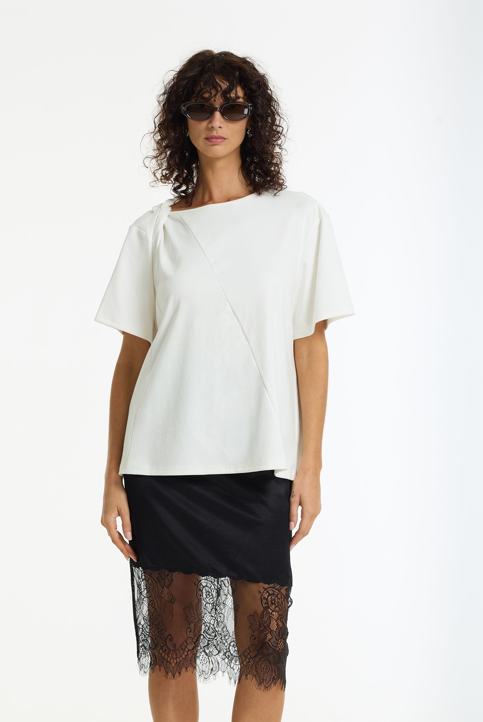 Woman wearing the Before Dawn Asymmetric Tee white cotton t-shirt by BLAK and black lace skirt on a white background