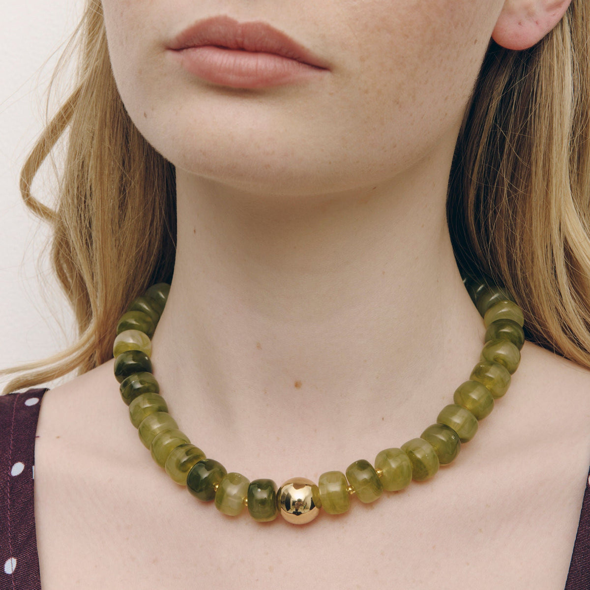 A close-up of a woman wearing a beaded necklace made of olive green translucent beads with a single polished gold accent bead at the center.