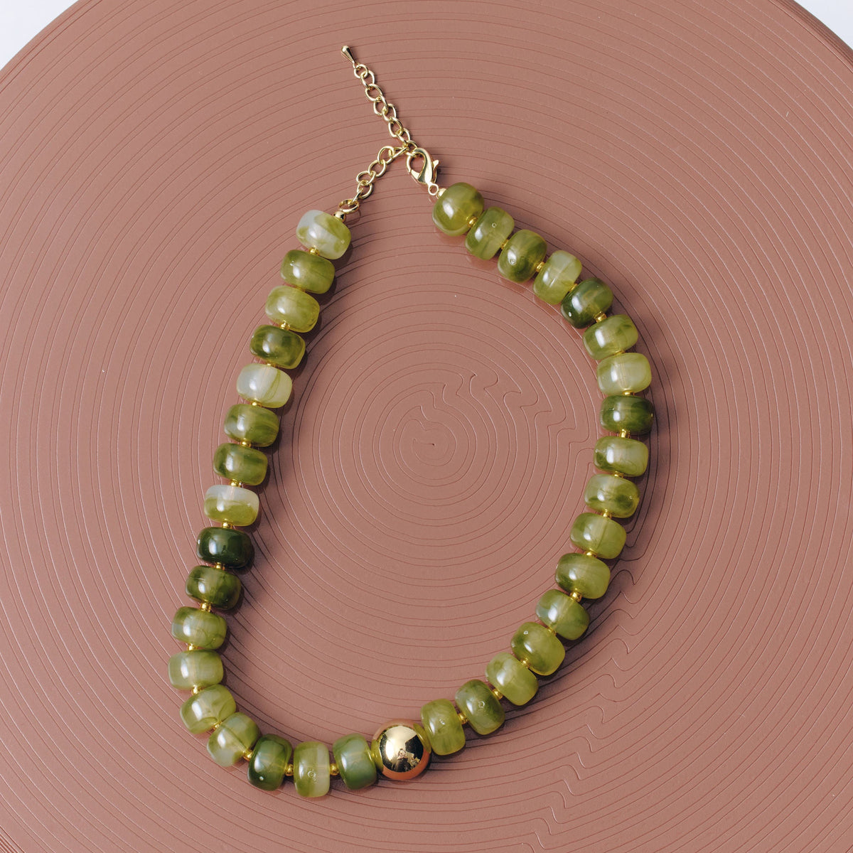 A beaded necklace made of polished green  beads with a small gold-toned accent bead and chain clasp, displayed on a round terracotta-colored surface.