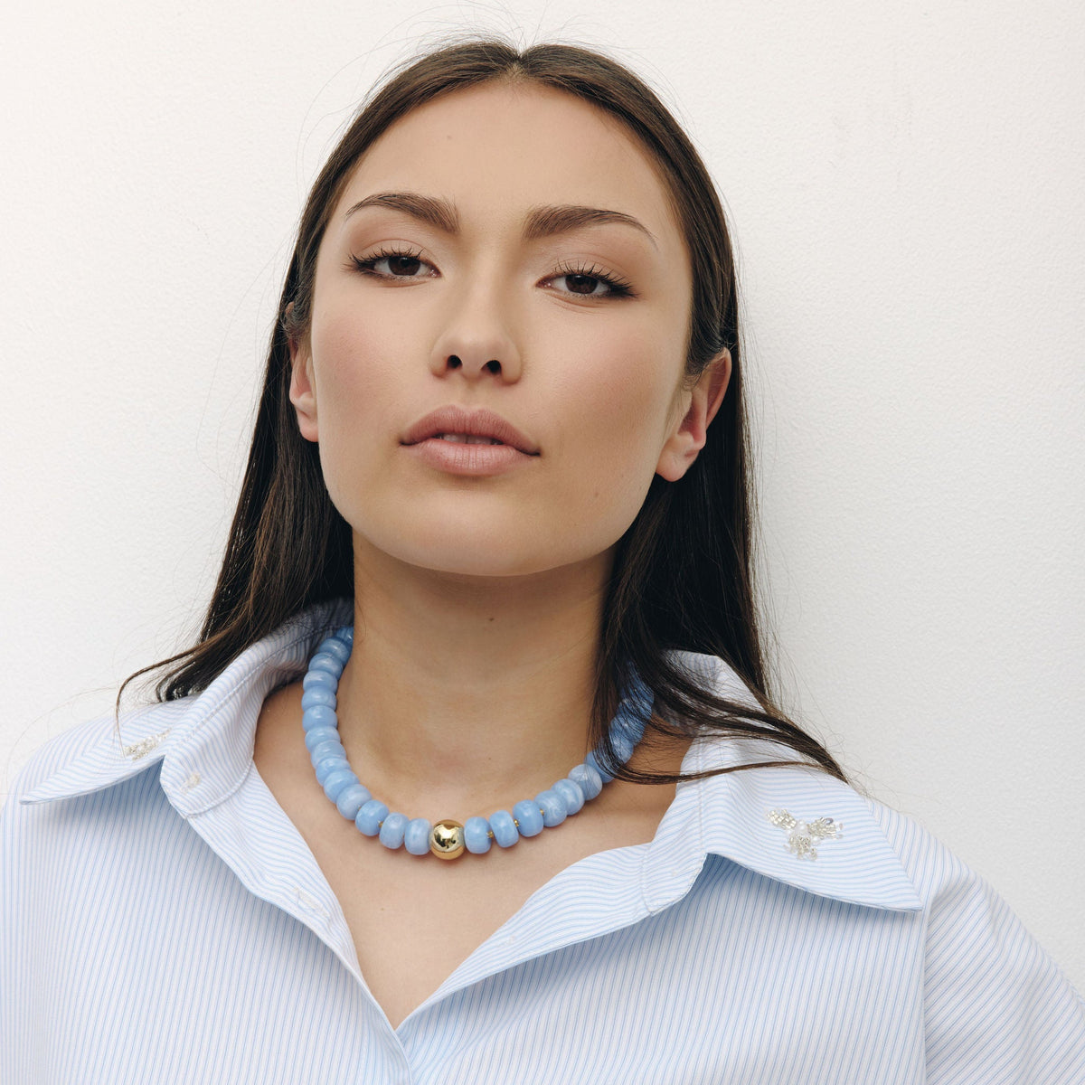 A woman wearing a light blue beaded necklace with a small gold accent bead, paired with a light blue and white striped button-down shirt, photographed against a plain off-white background.
