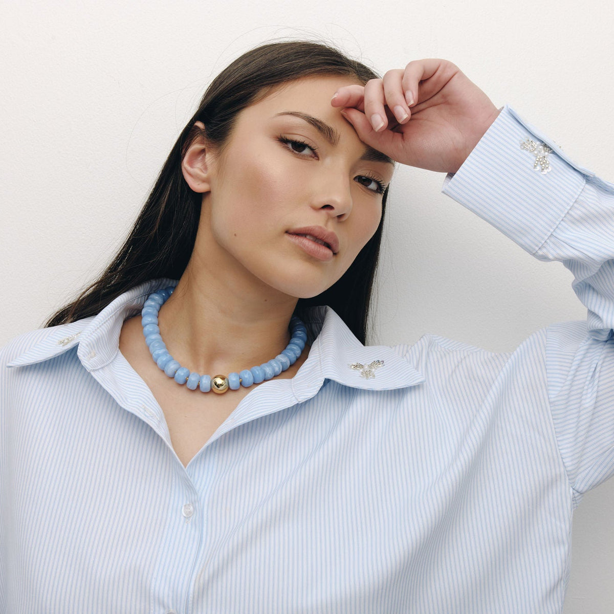 A woman with long dark hair wearing a light blue pinstriped button-down shirt and a light blue beaded necklace, posing against a plain off-white background.