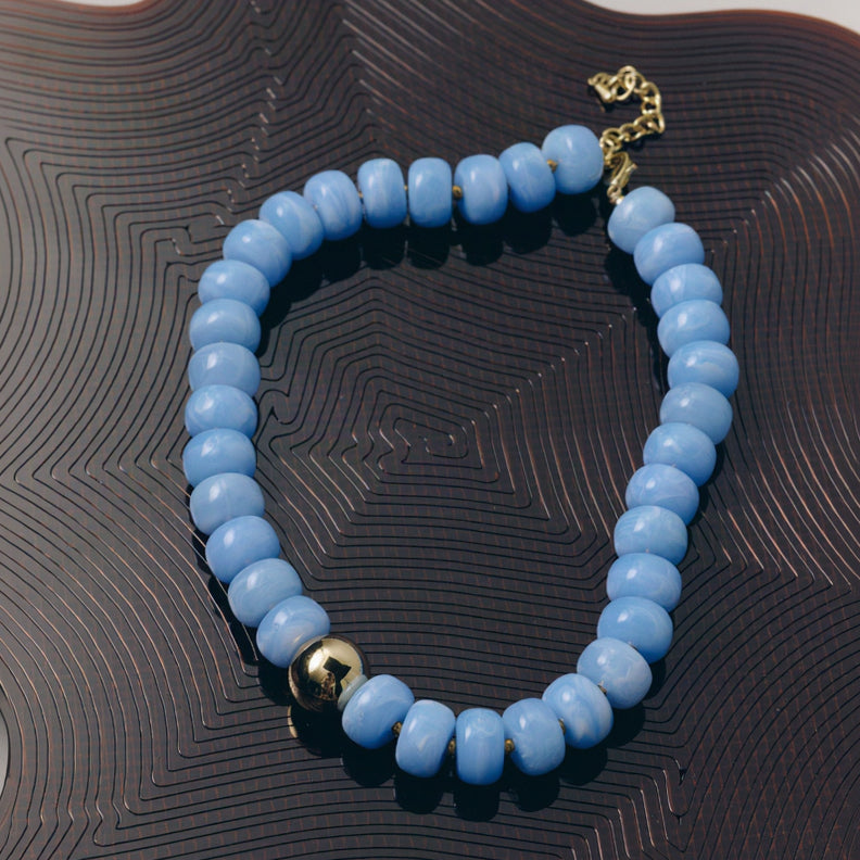 A beaded bracelet made of light blue cylindrical beads with a single polished gold accent bead and a gold clasp, displayed on a dark brown textured surface.