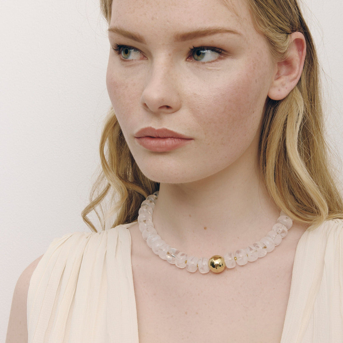 A close-up of a woman wearing a delicate pale pink beaded collar necklace with a central gold bead, paired with a cream-colored pleated top against a plain light background.