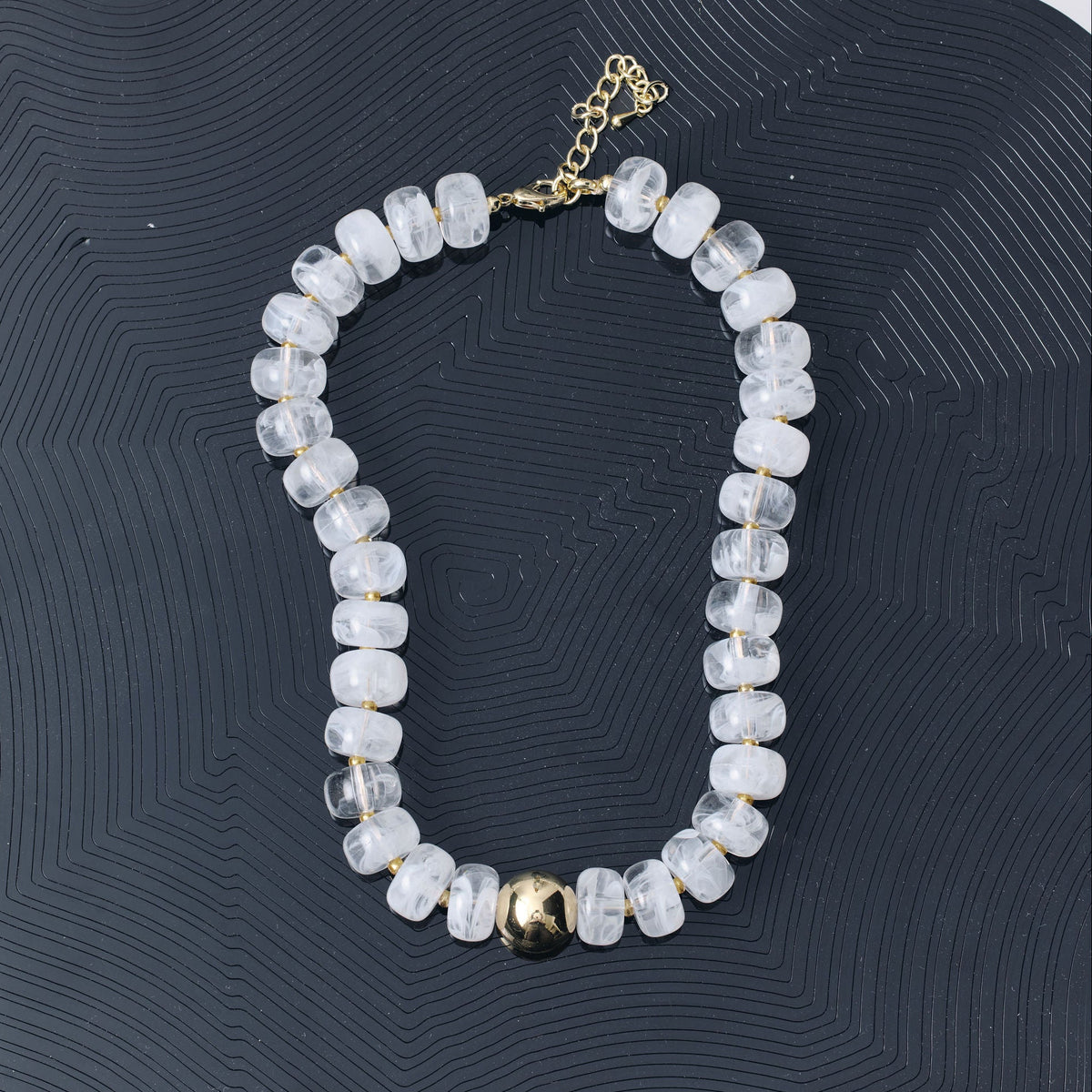 A beaded necklace made of clear faceted beads with a small gold ball accent and gold clasp, displayed on a dark wavy tray against a white background.