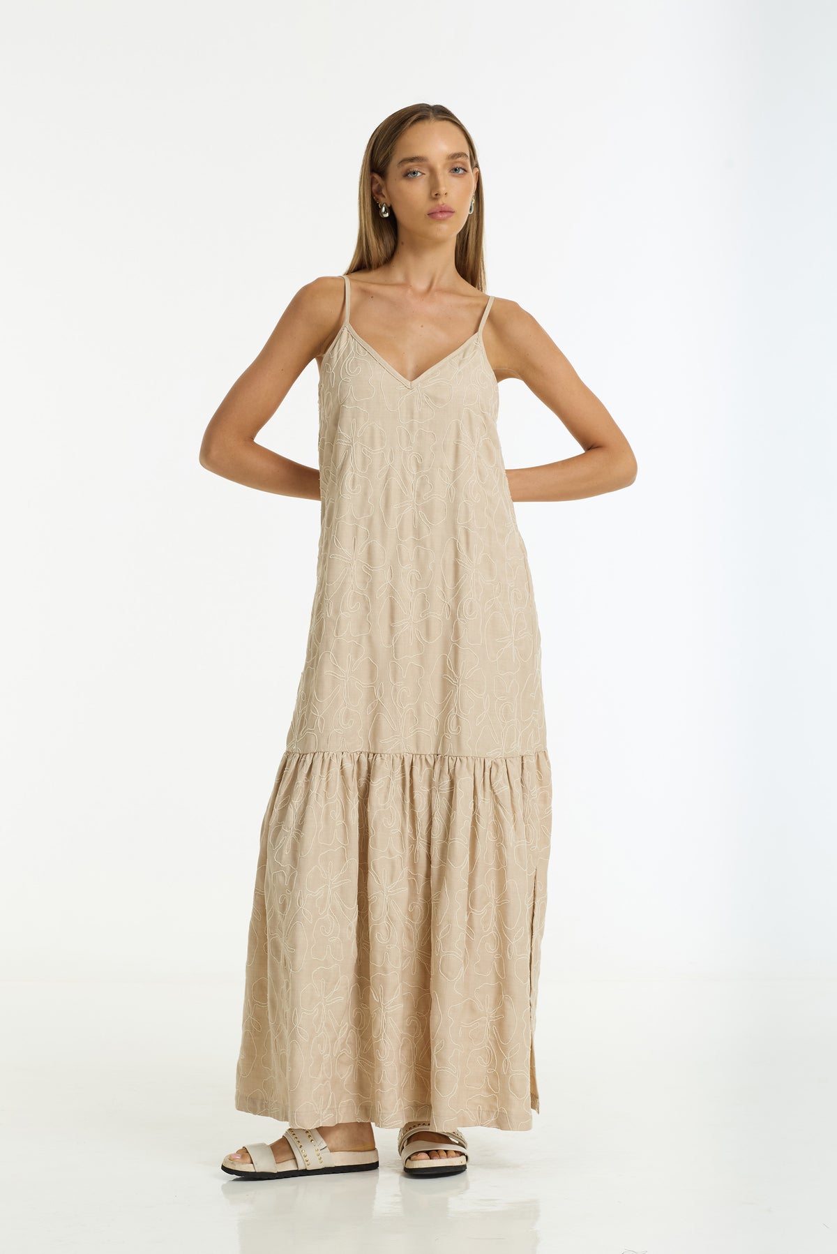 A woman modeling a Dallas Cotton Embroidered Maxi Dress in camel by BLAK, paired with beige slide sandals, against a plain white studio background.