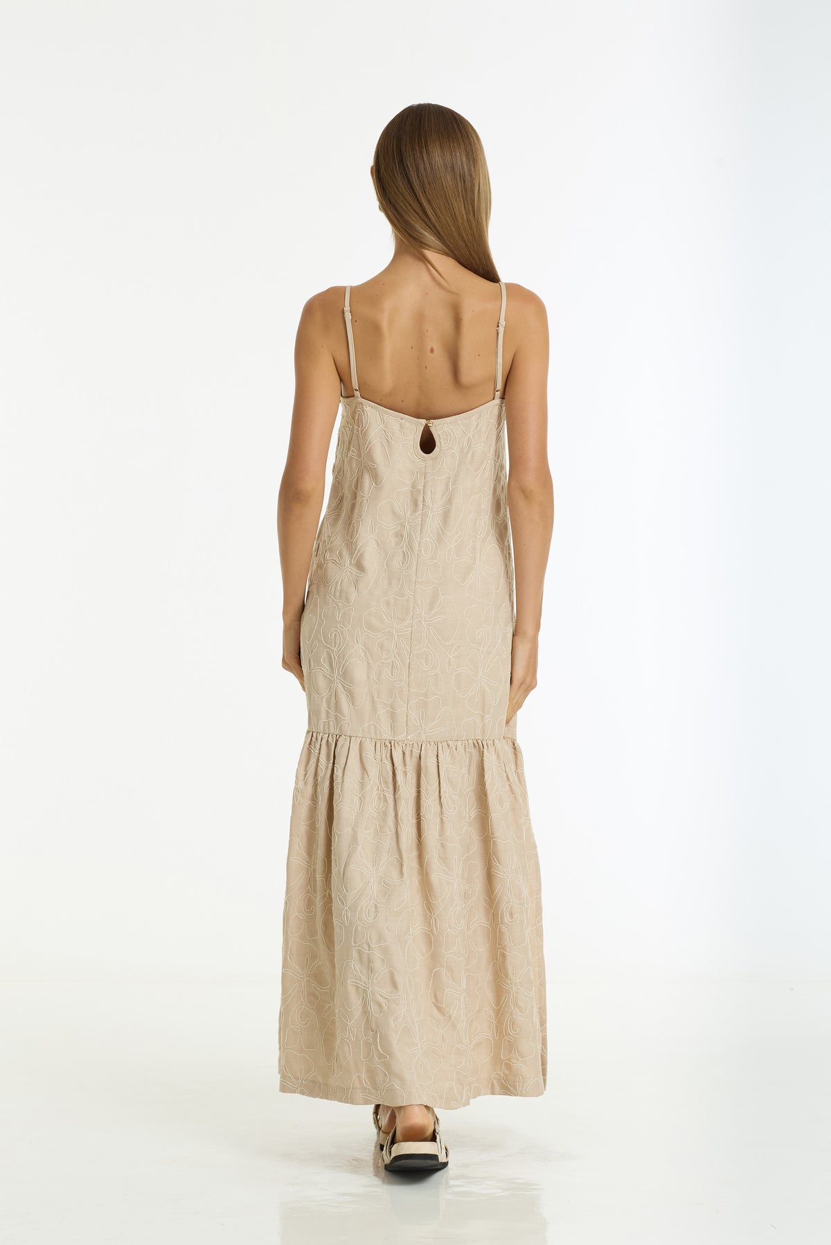 A woman viewed from behind wearing a Dallas Cotton Embroidered Maxi Dress in Camel by BLAK, standing against a plain white studio background.