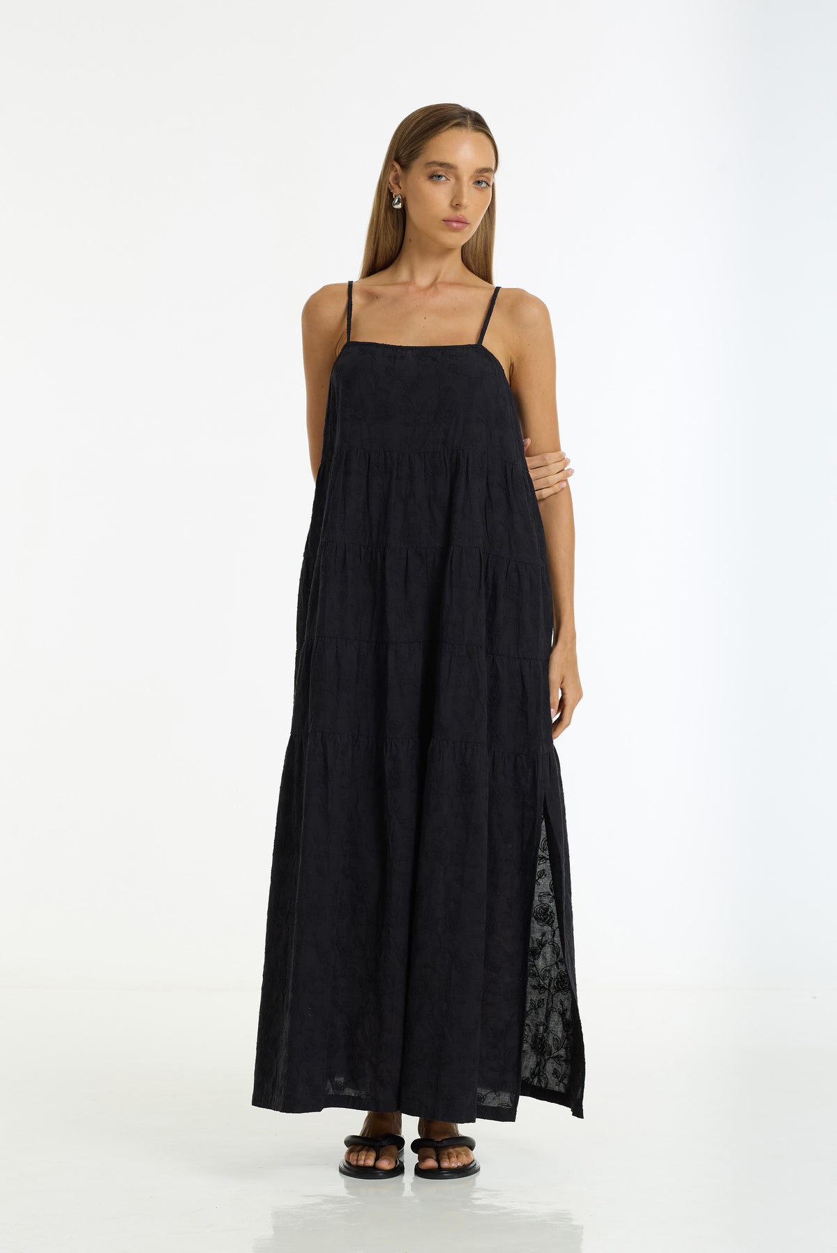 A woman modeling a black maxi dress with thin spaghetti straps, a square neckline, and a side slit showing a sheer black lace lining, set against a plain white studio background.