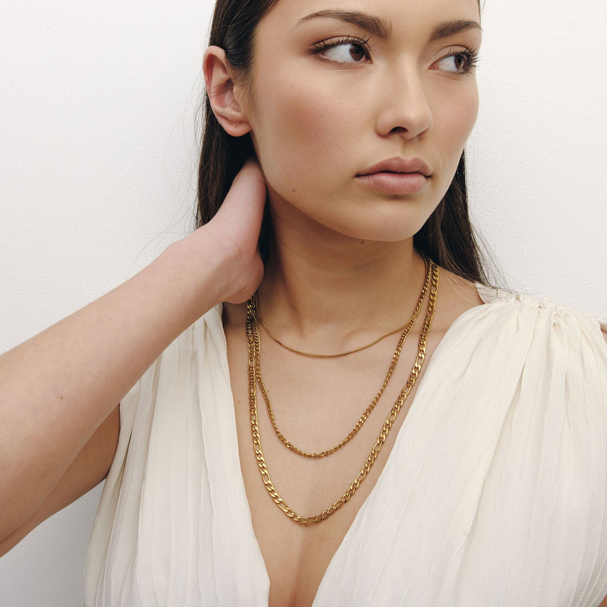 A woman wearing layered gold necklaces, including a chain link necklace and a delicate chain necklace, paired with a white sleeveless top, set against a plain light background.