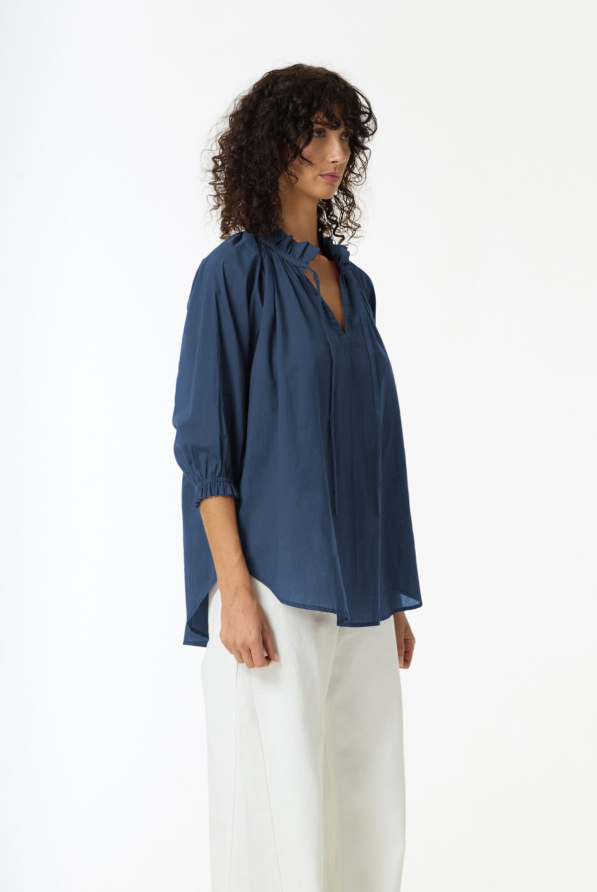 A woman with curly dark hair models a navy blue blouse with a split neckline and white trousers against a plain white studio background.