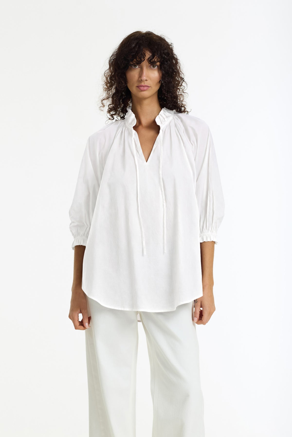 A model with curly dark hair wearing a white long-sleeve blouse with a split V-neck and gathered sleeves, paired with white trousers, against a plain white studio background.
