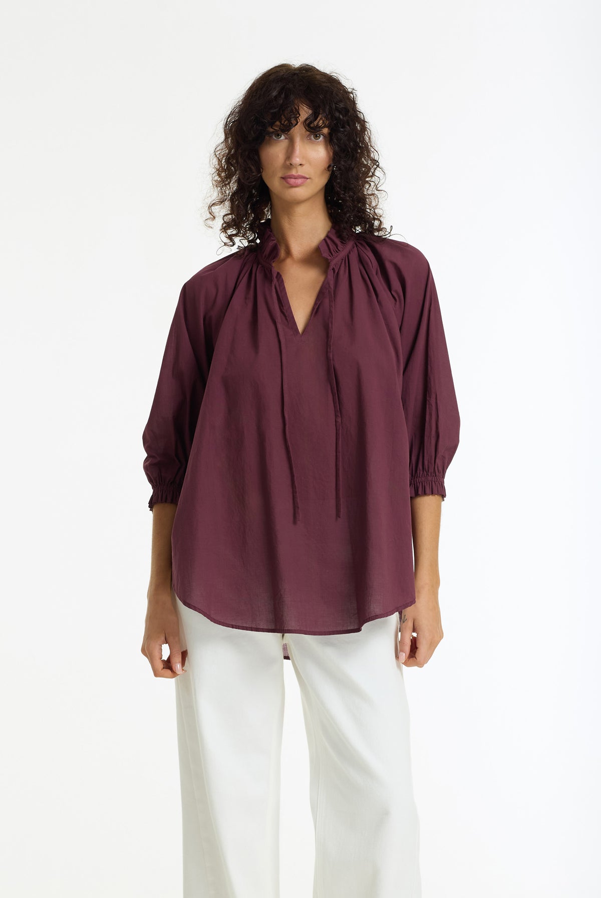 A woman modeling a Charlotte Cotton Voile Top in Winter Wine by BLAK, paired with white trousers, against a plain white studio background.