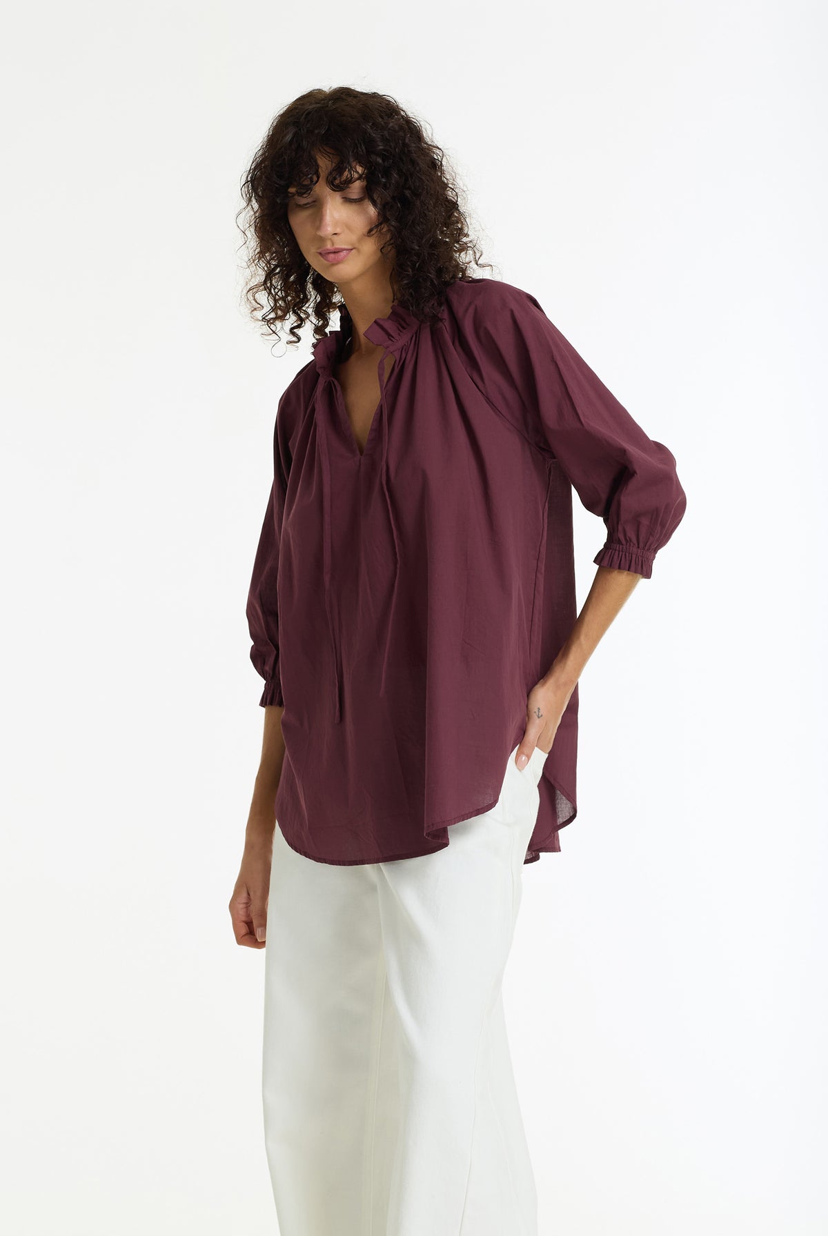 A woman modeling a loose-fitting, deep plum-colored blouse with a split neckline and gathered sleeves, paired with white trousers, against a plain white studio background.