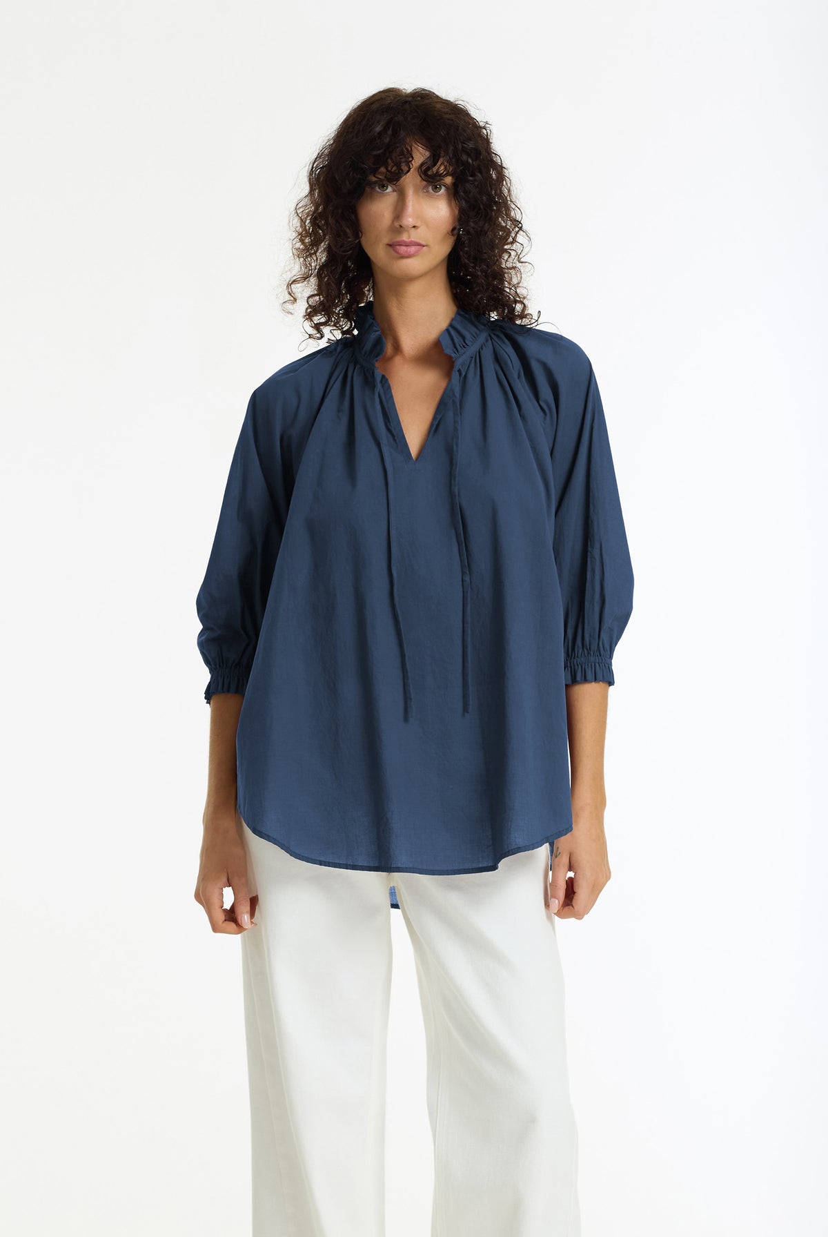 A woman modeling a navy blue long-sleeve blouse with a gathered, tie-front neckline and white pants, photographed against a clean white studio background.
