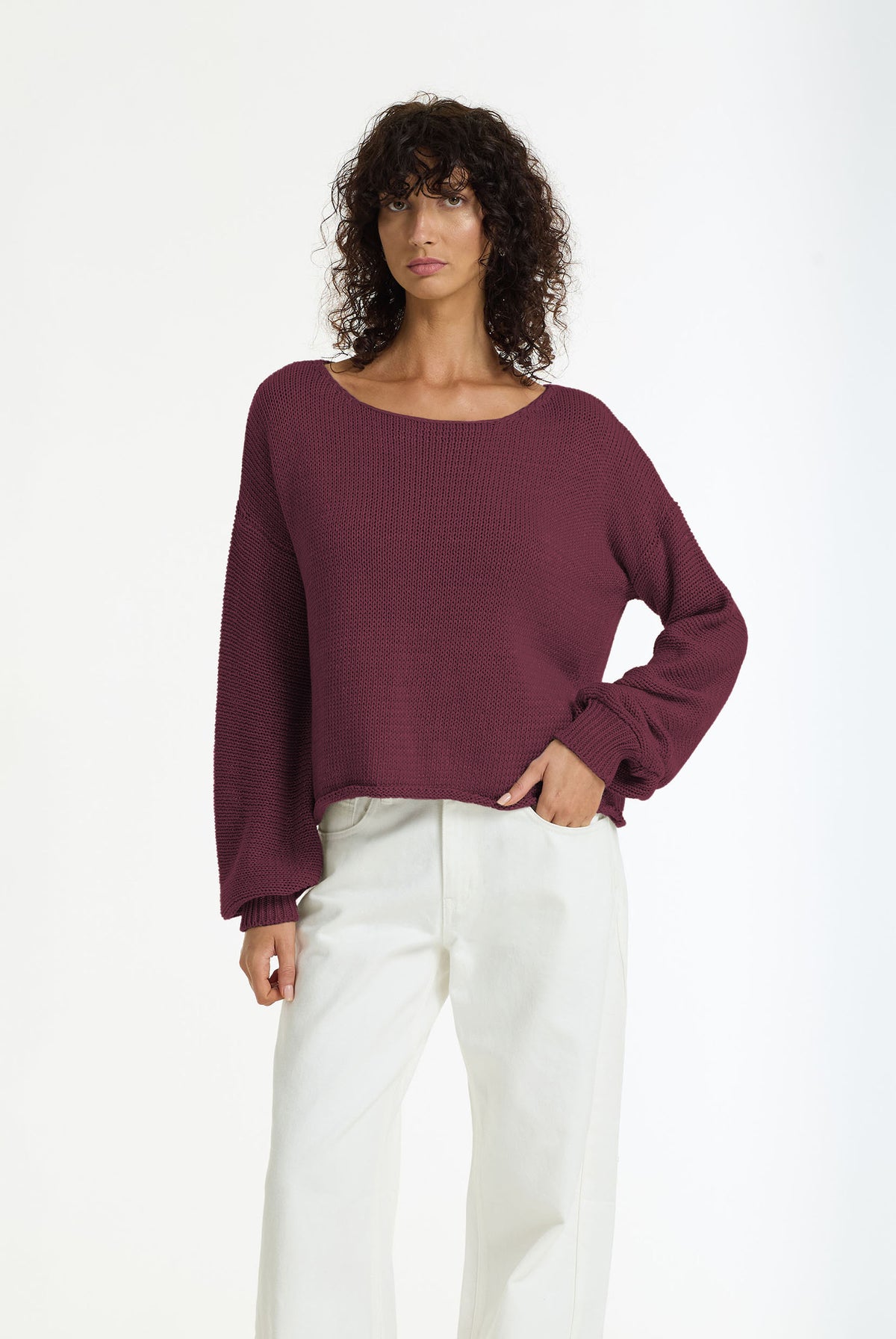 A woman modeling a Close Cotton Yarn Sweater in Winter Wine by BLAK, paired with white trousers, against a plain white studio background.