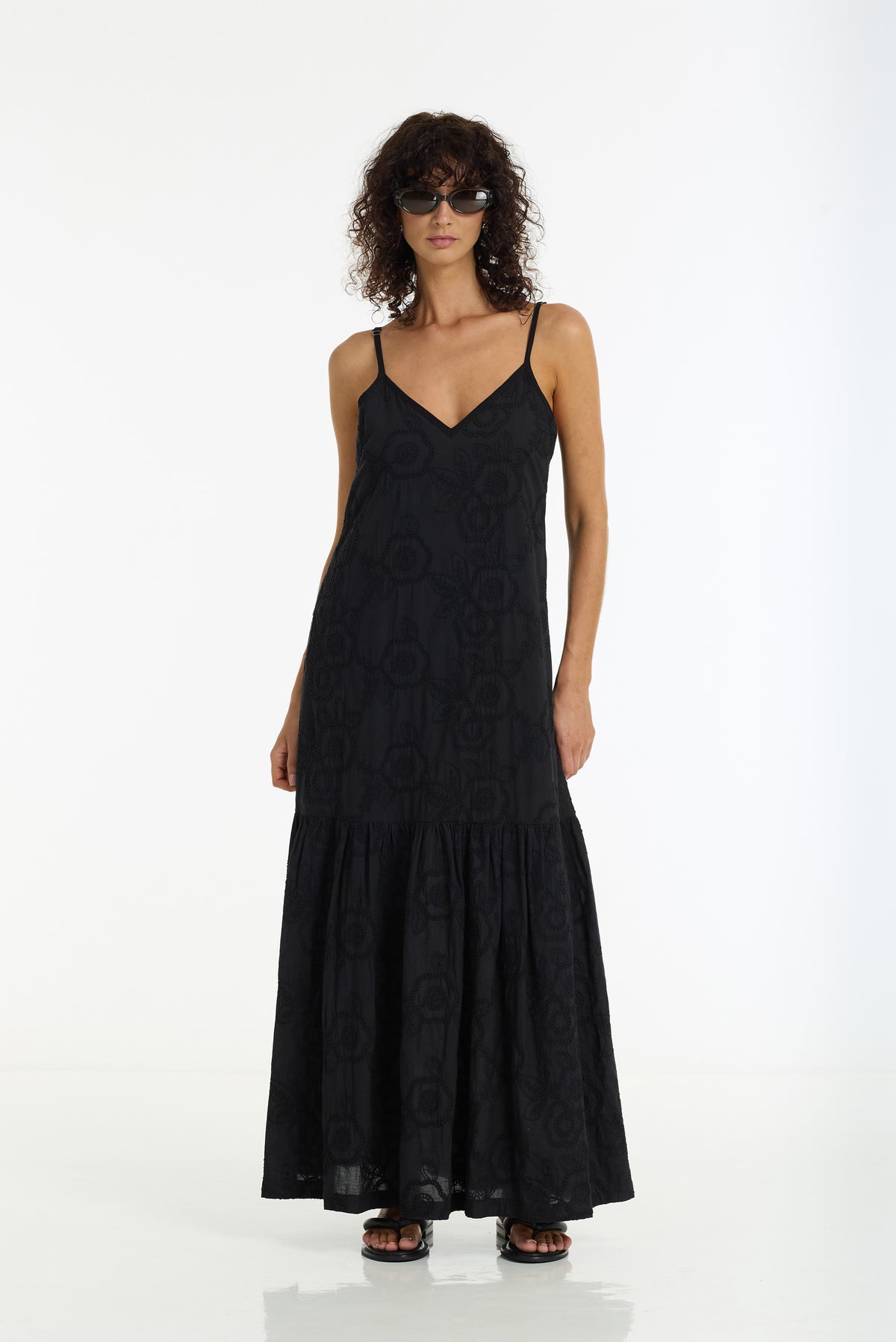 A woman modeling a black maxi dress with thin spaghetti straps and a subtle tonal circular jacquard pattern, photographed against a seamless white studio background.