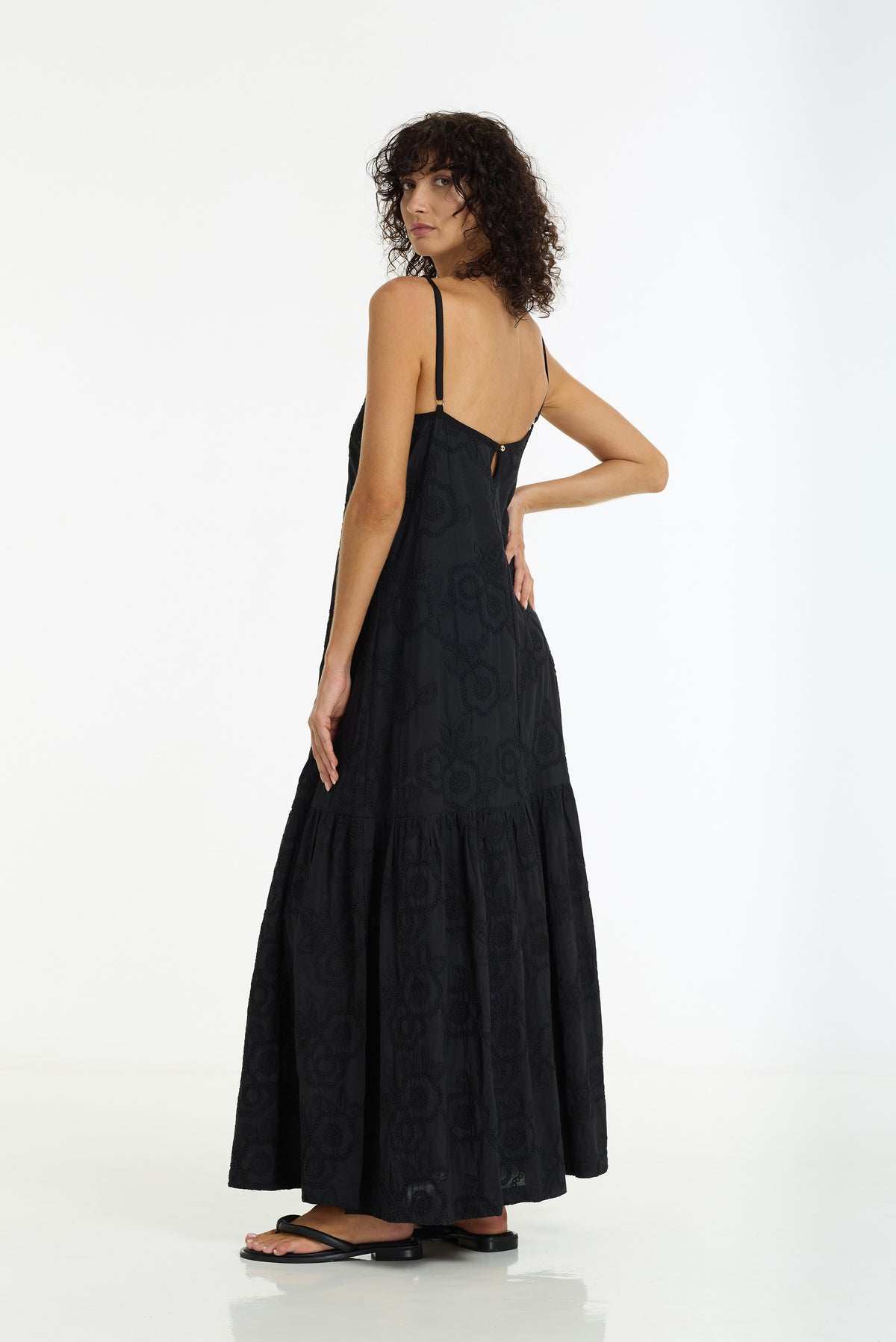 A woman modeling a black maxi dress with thin spaghetti straps, a backless open back, and a tiered ruffled skirt, shown from a three-quarters back view against a white studio background.