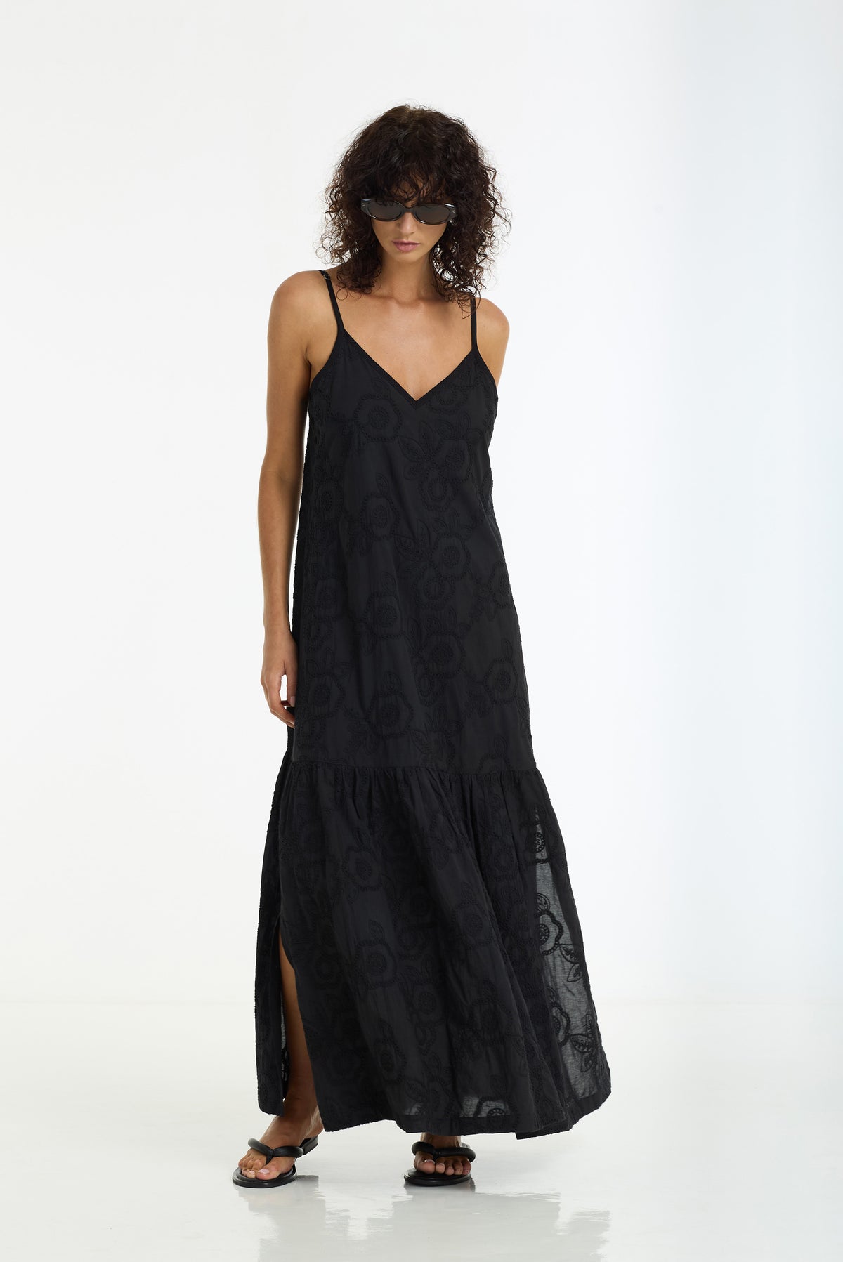 A woman modeling a black maxi dress with thin straps, a V-neckline, and a tiered skirt with a side slit, photographed against a plain white studio background.