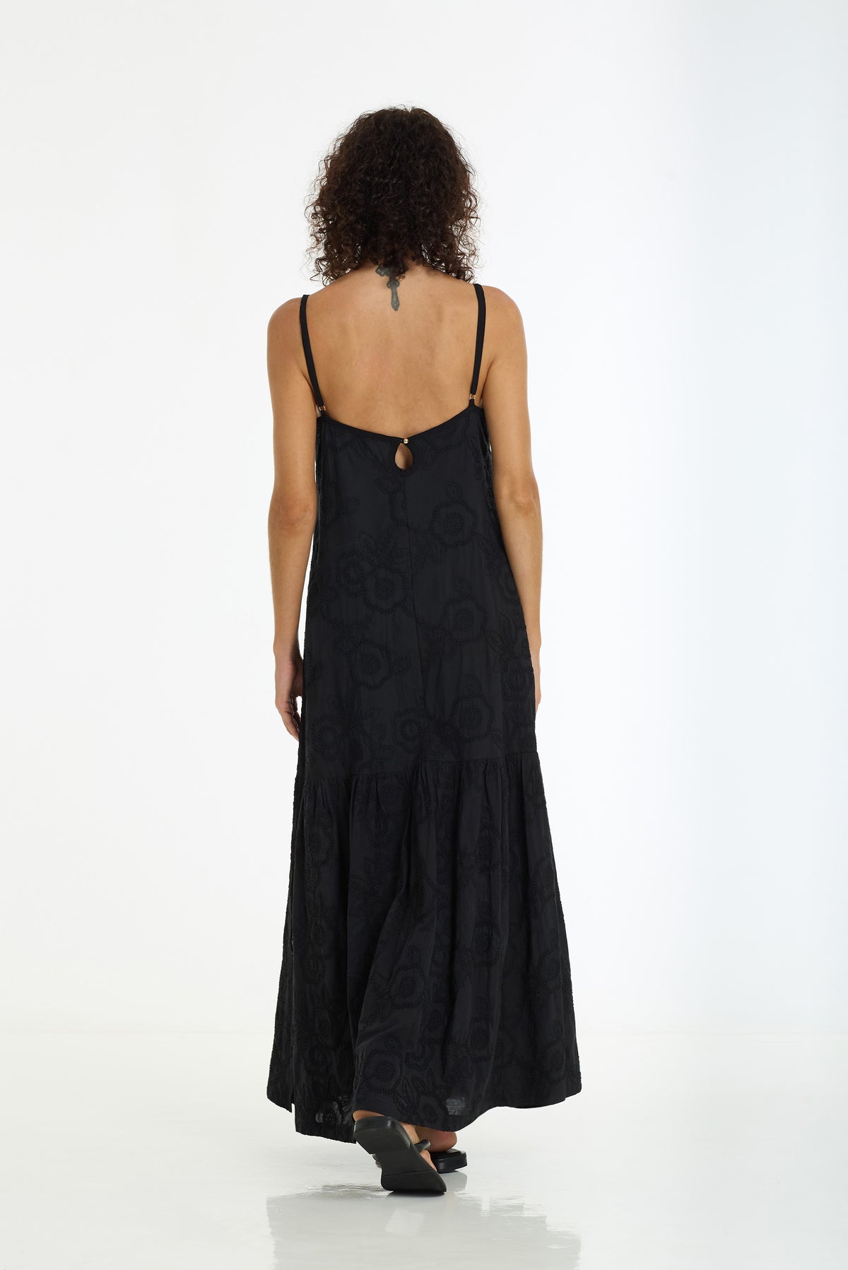 A woman viewed from behind wearing a black spaghetti-strap maxi dress with a subtle tonal circular pattern and a small gold keyhole detail at the back neckline, set against a plain white studio background.