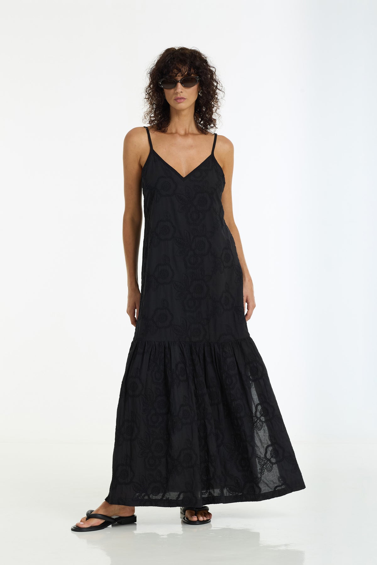 A model wearing a Dallas Cotton Anglaise Maxi Dress in Black by BLAK, styled with black flat sandals against a plain white studio background.