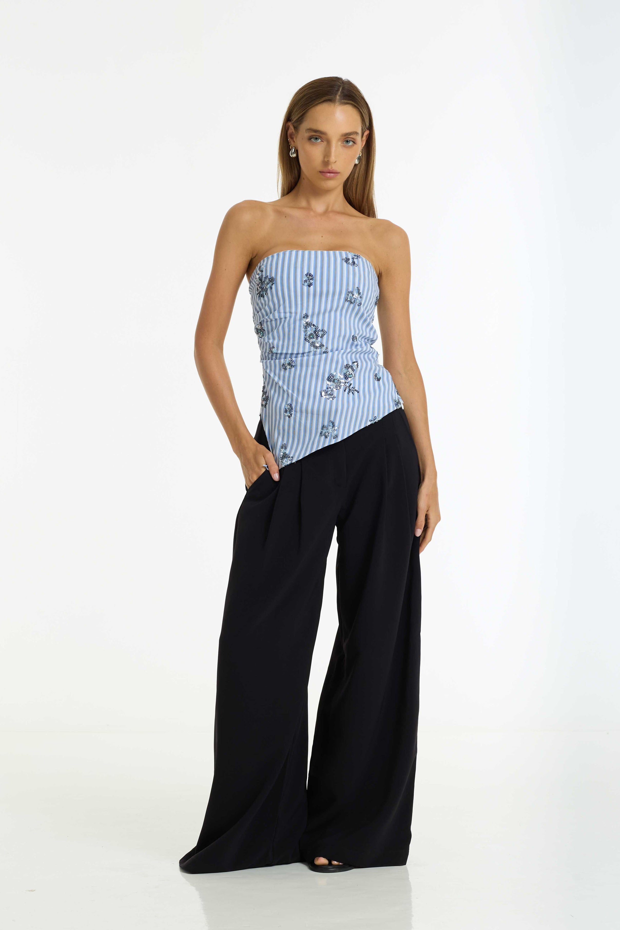 A woman modeling a strapless top with a light blue and white vertical stripe pattern and small dark floral motifs, paired with the Dante Wide Leg Pant in black by BLAK, against a plain white studio background.