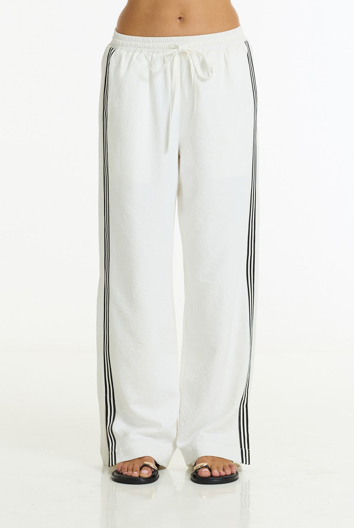 White wide-leg sweatpants with black side stripes, modeled from the waist down with black sandals on a white studio background.