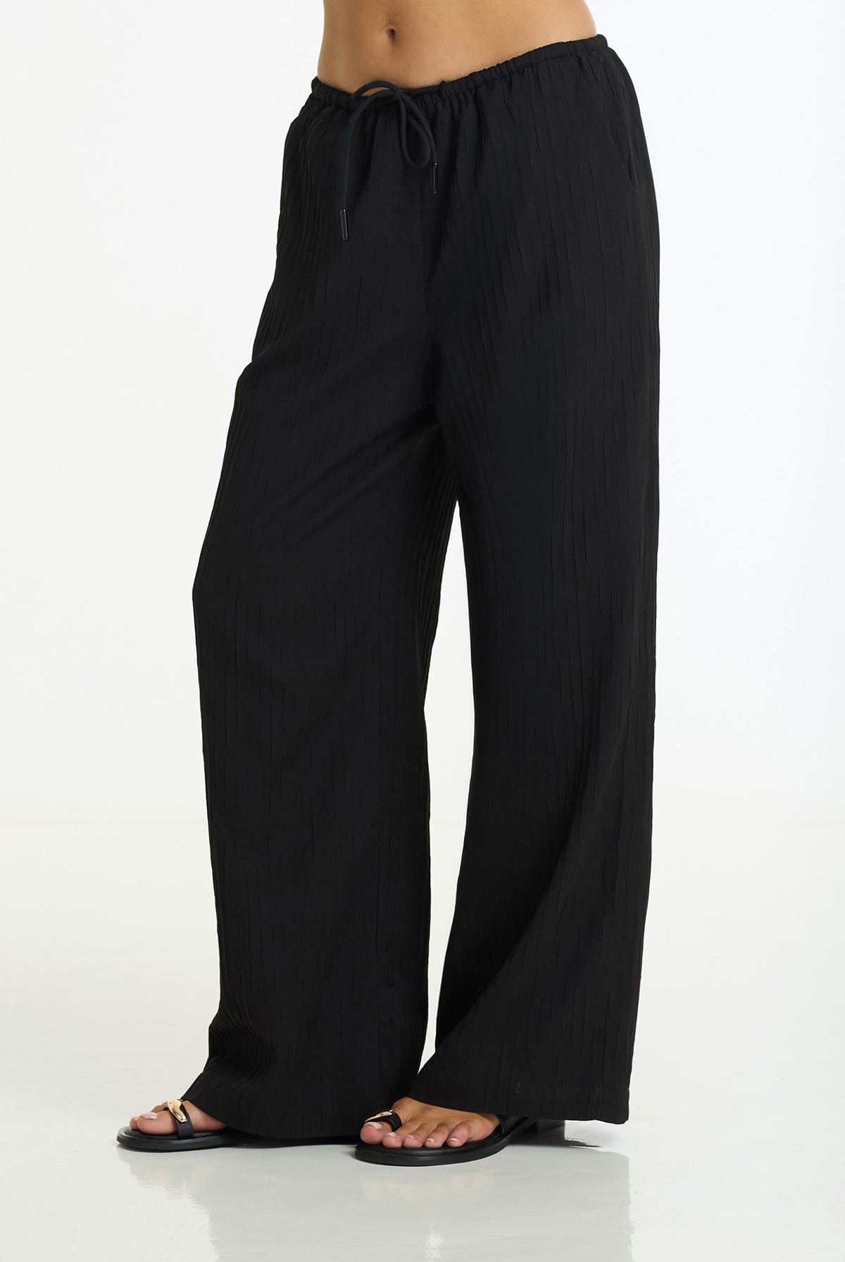 Black wide-leg drawstring pants with a subtle vertical crinkle texture, shown on a model wearing black sandals against a white studio background.