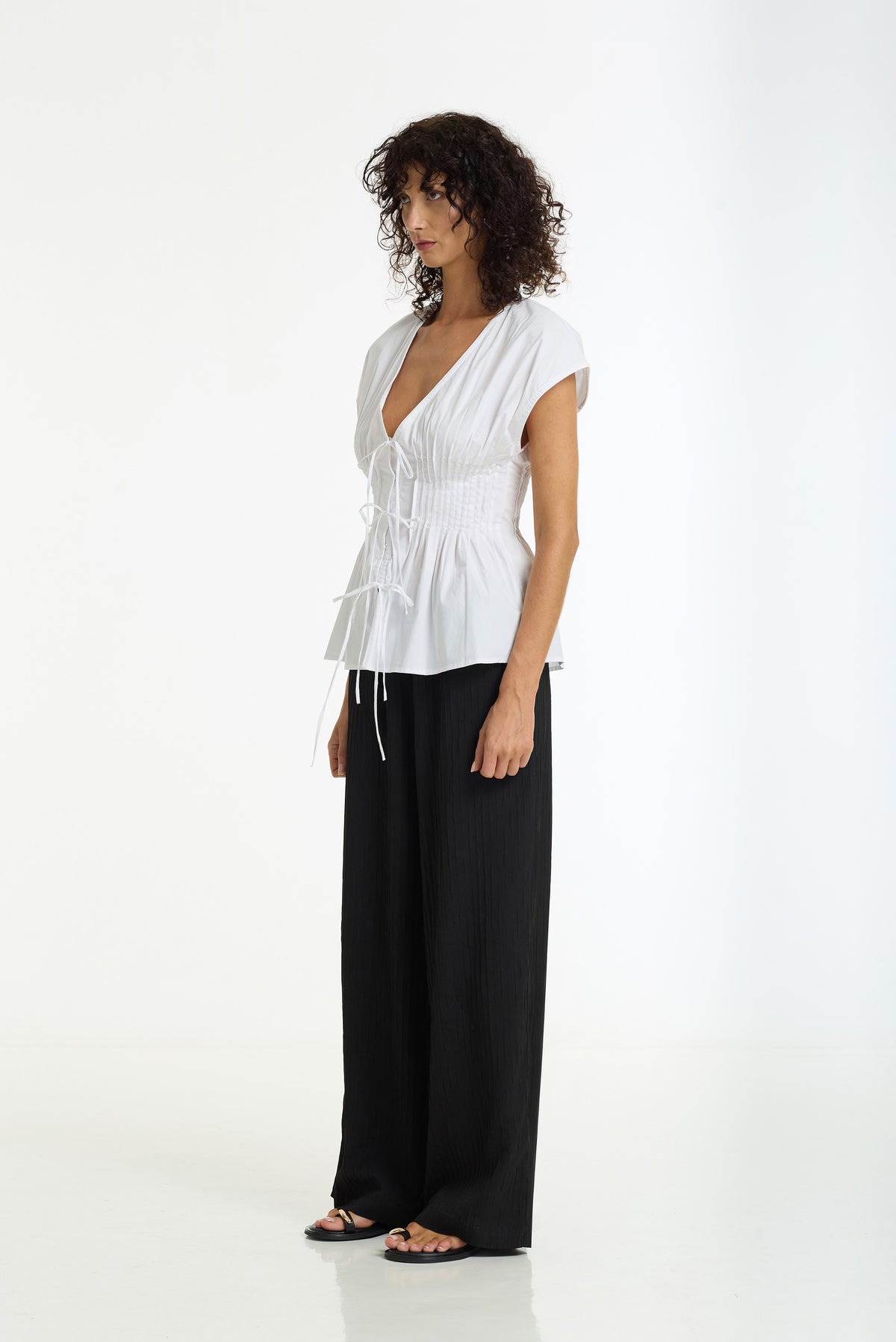 A woman stands in profile wearing a white short-sleeve smocked peplum top with a drawstring and black wide-leg pants against a clean white studio background.