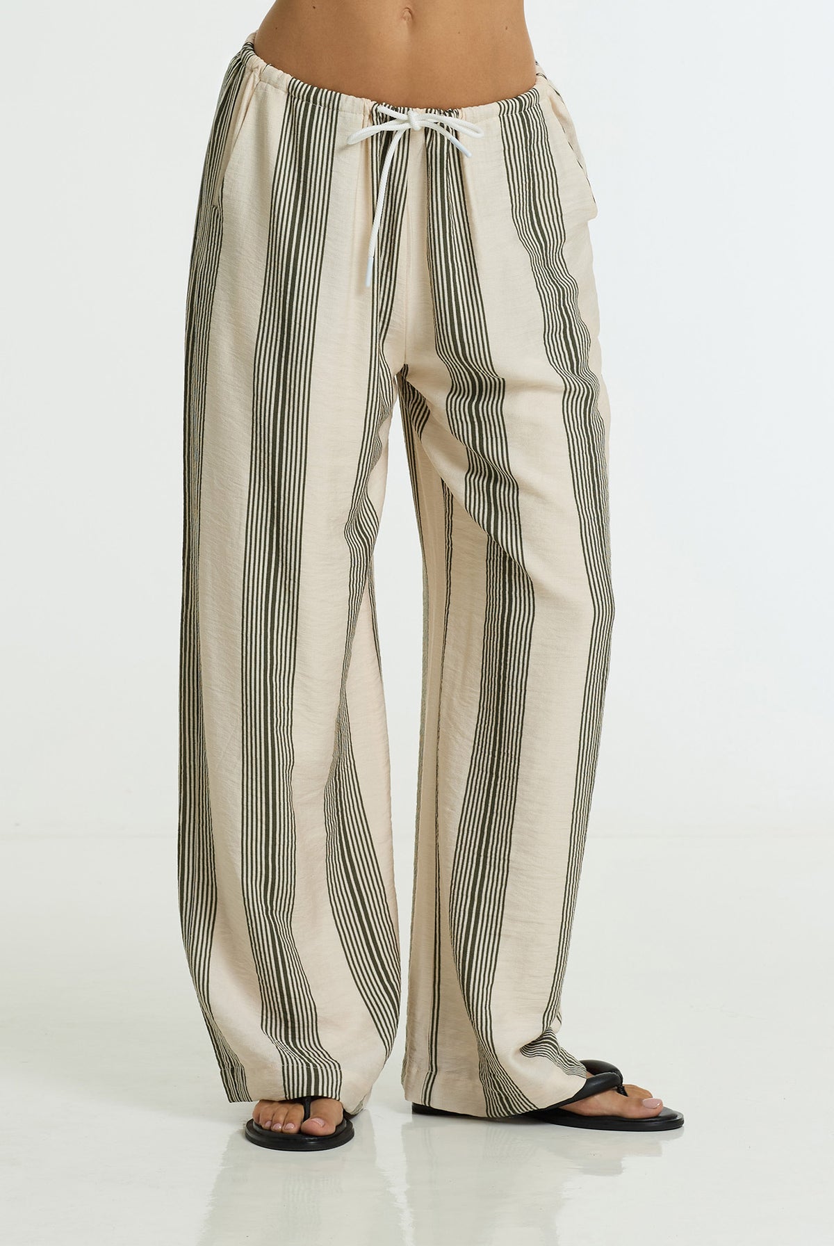 A person wearing loose, wide-leg drawstring pants with vertical khaki stripes on a cream base, paired with black flip-flop sandals, shown against a plain white studio background.