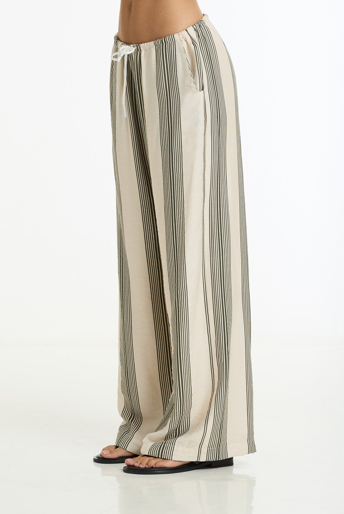 A person wearing loose, wide-leg striped pants with a drawstring waist and black flat sandals, shown from the waist down against a clean white studio background.