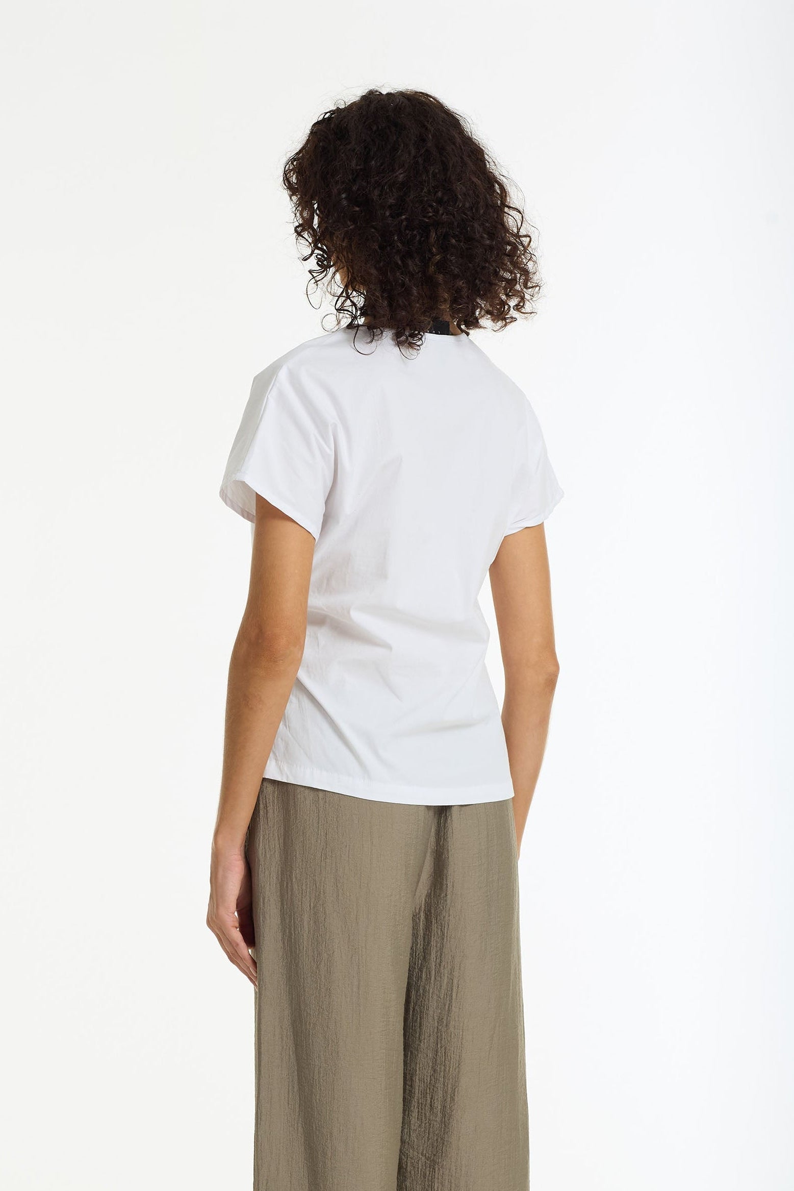 A woman with curly dark hair is shown from the back wearing a plain white short-sleeve t-shirt and light brown wide-leg trousers against a clean white studio background.