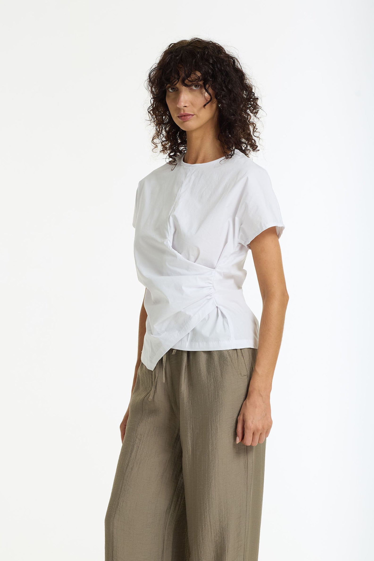 A model stands against a plain white studio background wearing a Endless Devotion Structured Cotton Top in white by BLAK and wide-leg taupe pants.