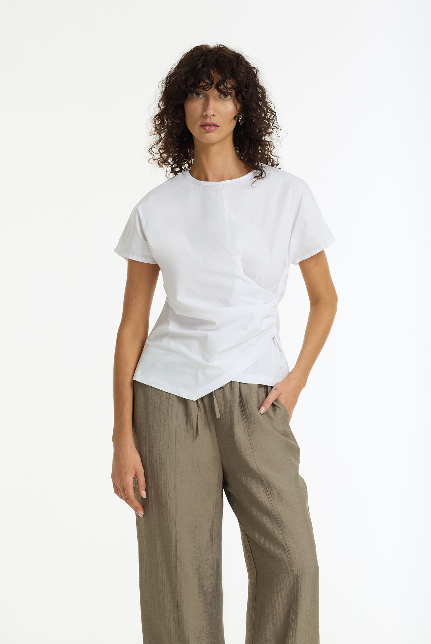 A woman modeling a Endless Devotion Structured Cotton Top in white by BLAK wih an asymmetrical hem and olive green wide-leg trousers, shot against a plain white studio background.