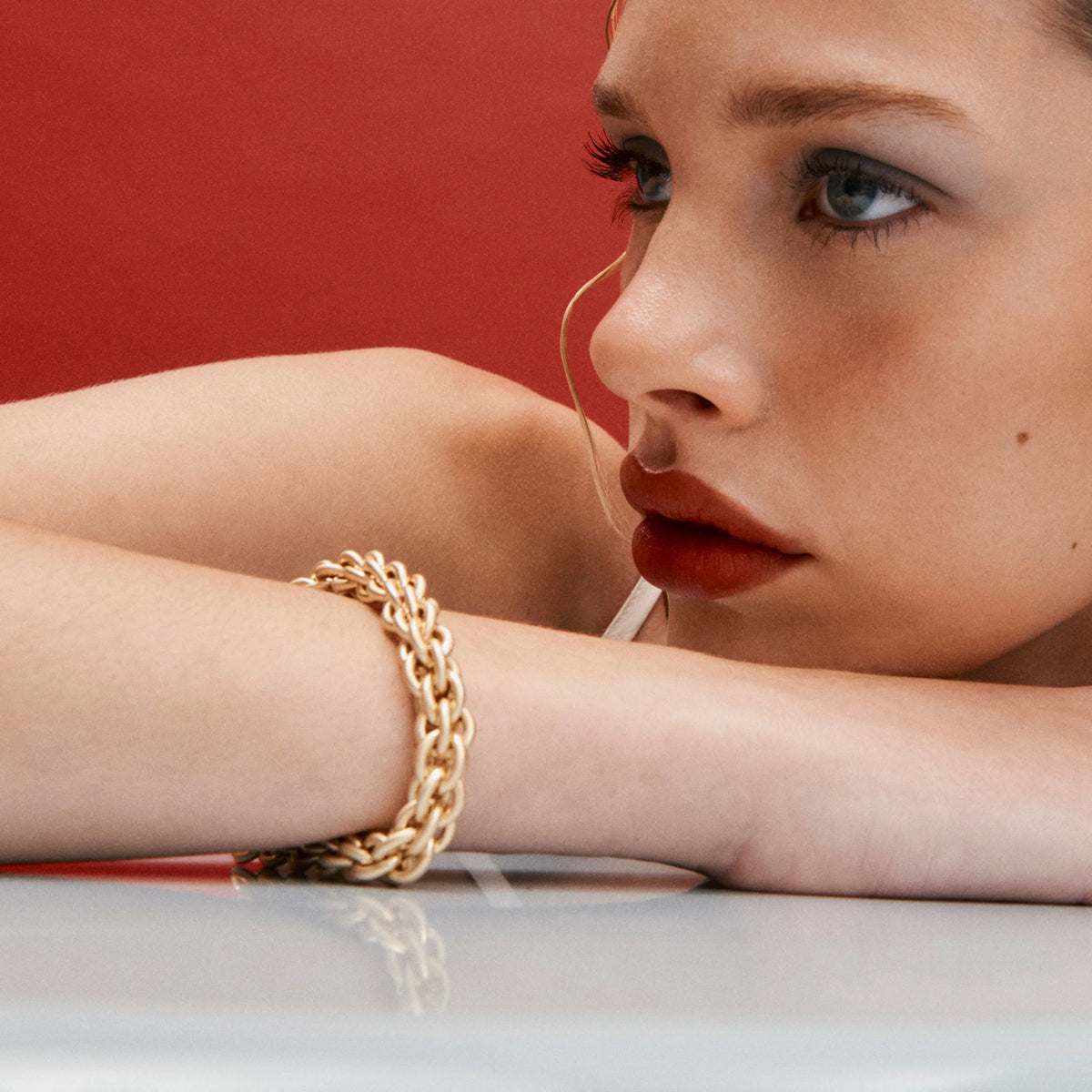 A close-up lifestyle shot of a model resting her head on a white surface, wearing a chunky gold chain link bracelet against a solid red background.