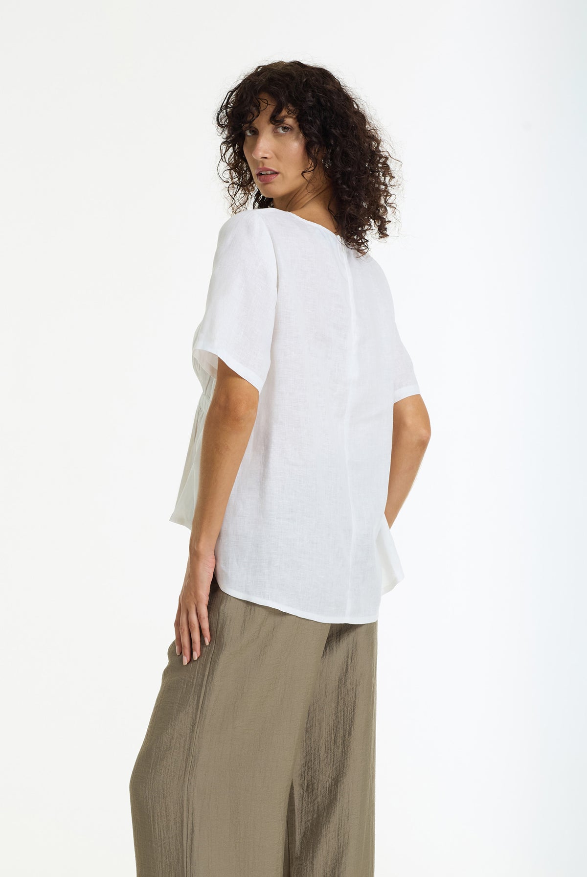 A woman with curly dark hair is shown from a back-three-quarter angle wearing a loose white short-sleeve linen top and olive green wide-leg pants against a plain white studio background.