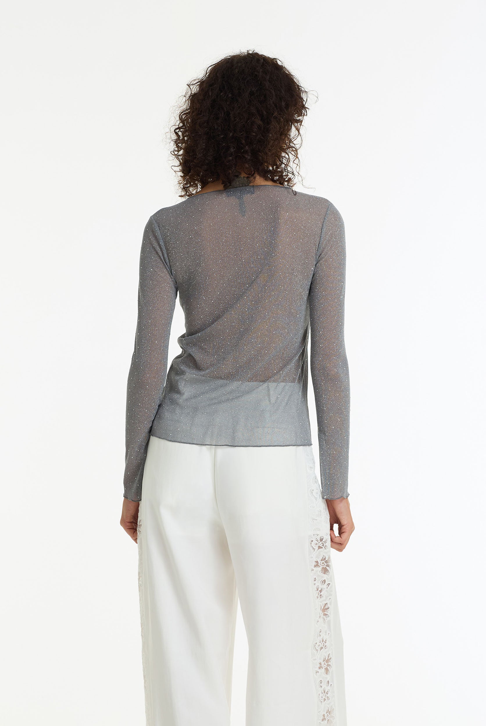 A back view of a person wearing the Gigi Diamanté Long Sleeve Top by BLAK, a charcoal sheer stretch top with silver diamantés, long sleeves and a softly fitted silhouette and white pants on a white background.