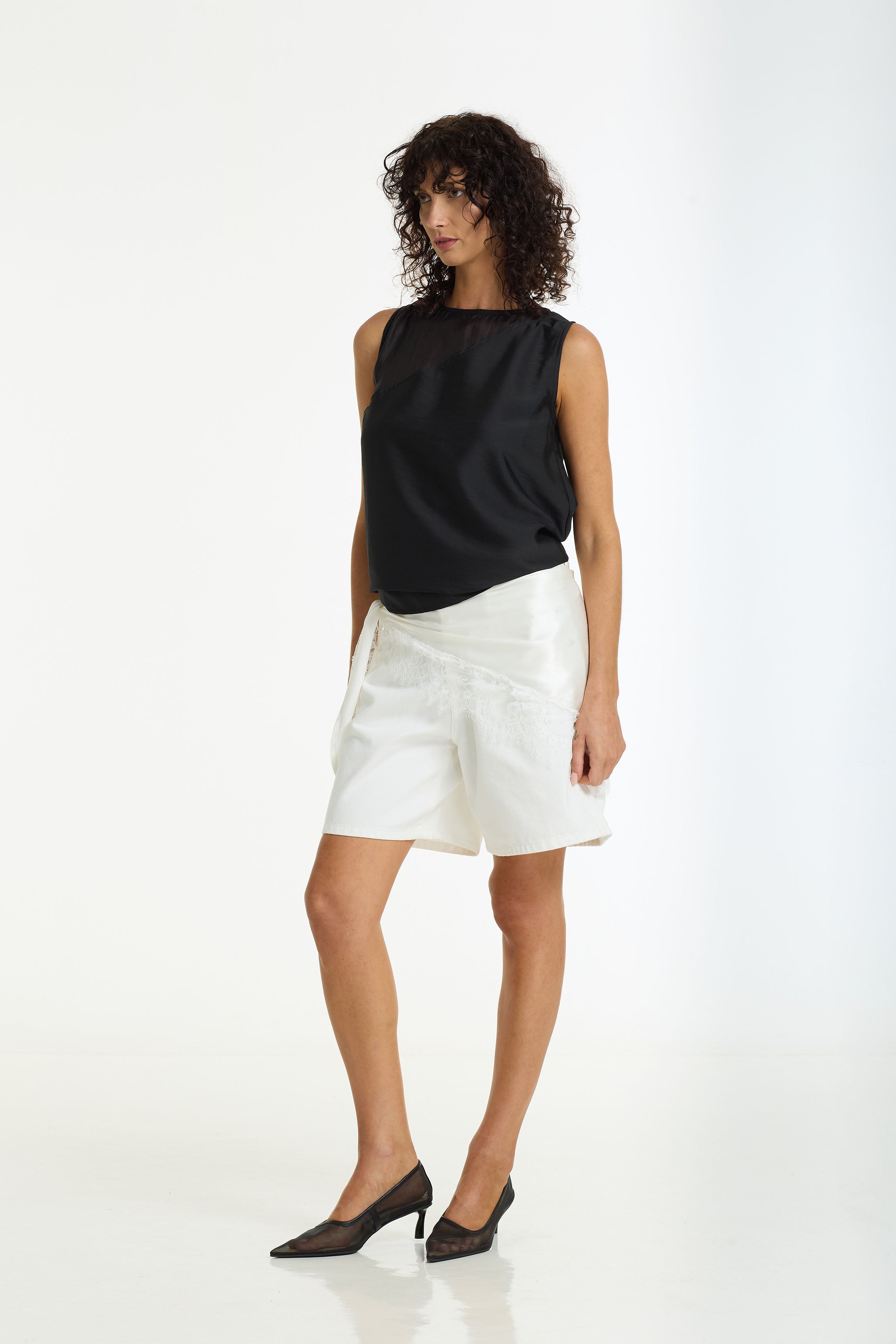 A model stands against a white studio background wearing a sleeveless black top with a sheer yoke and white shorts, paired with The Lace Edit Bandana Scarf in ivory by BLAK.